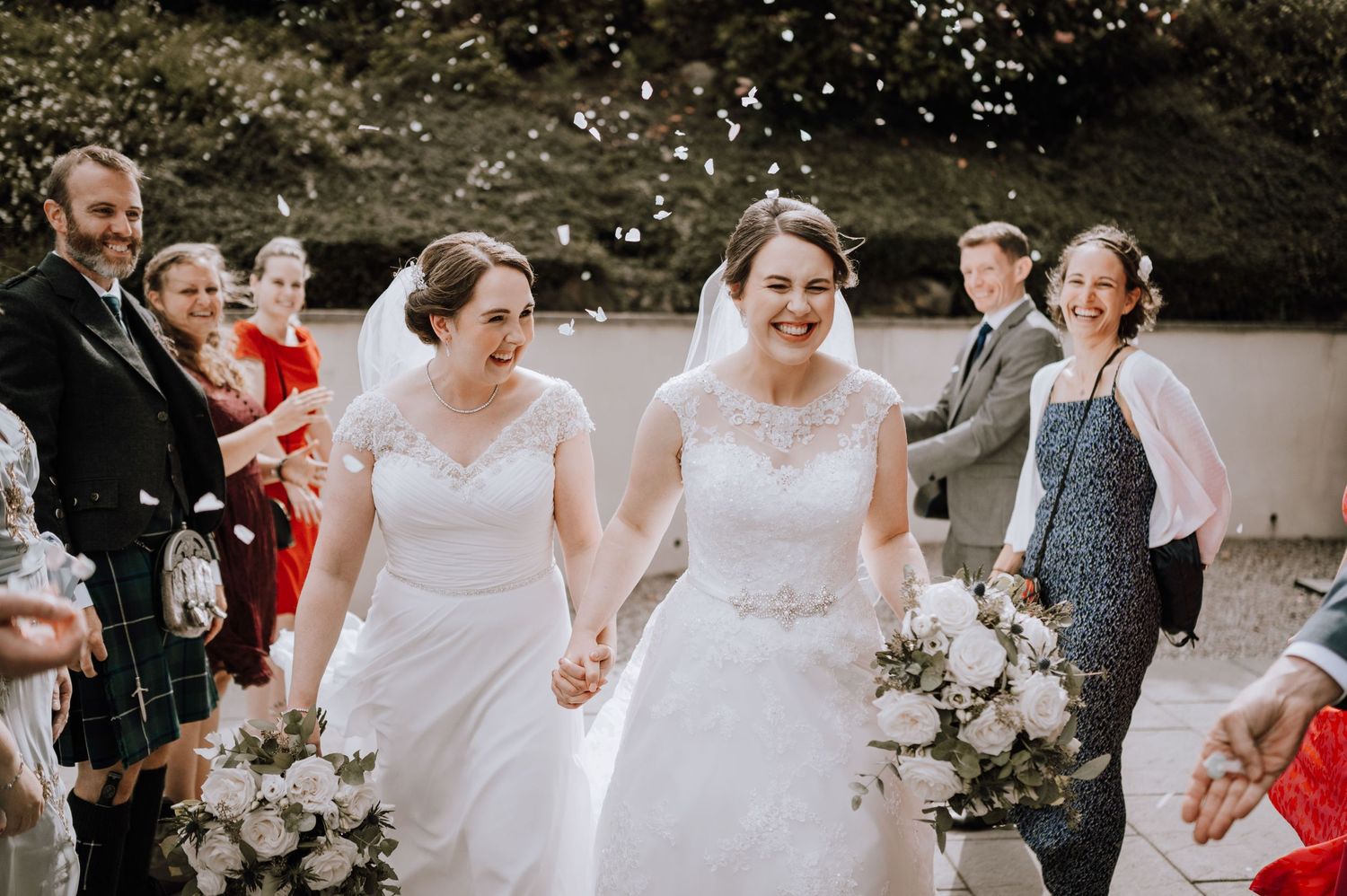 Two people in wedding dresses walk together amid confetti and celebrating guests outdoors.