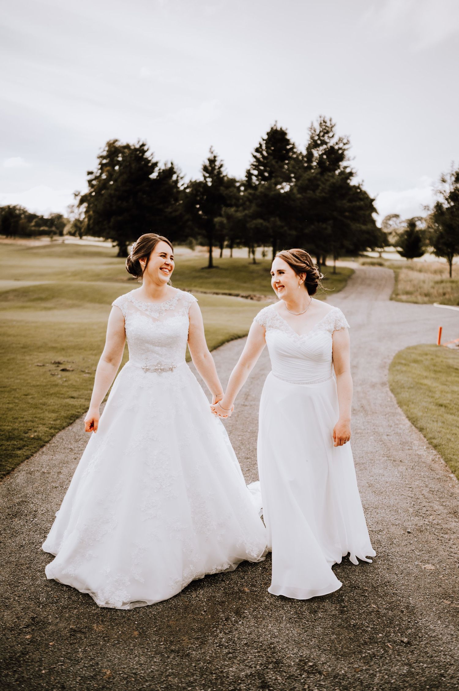 Couple in white wedding dresses walking together on a path near a golf course.