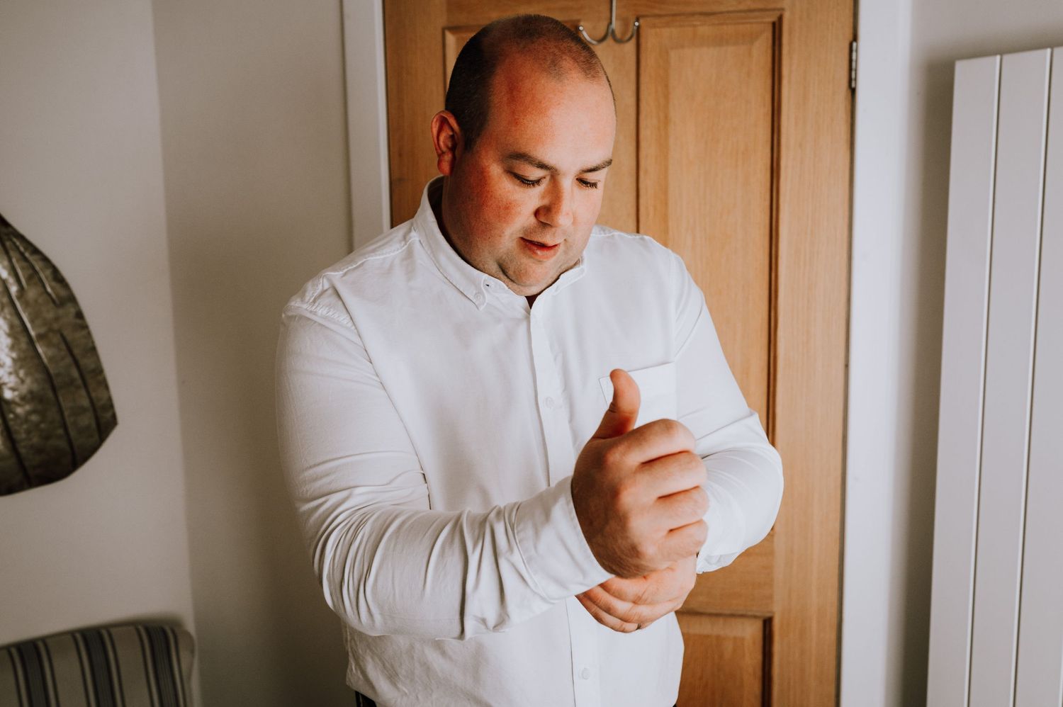 A person in white buttoning their shirt cuffs by a wooden door.