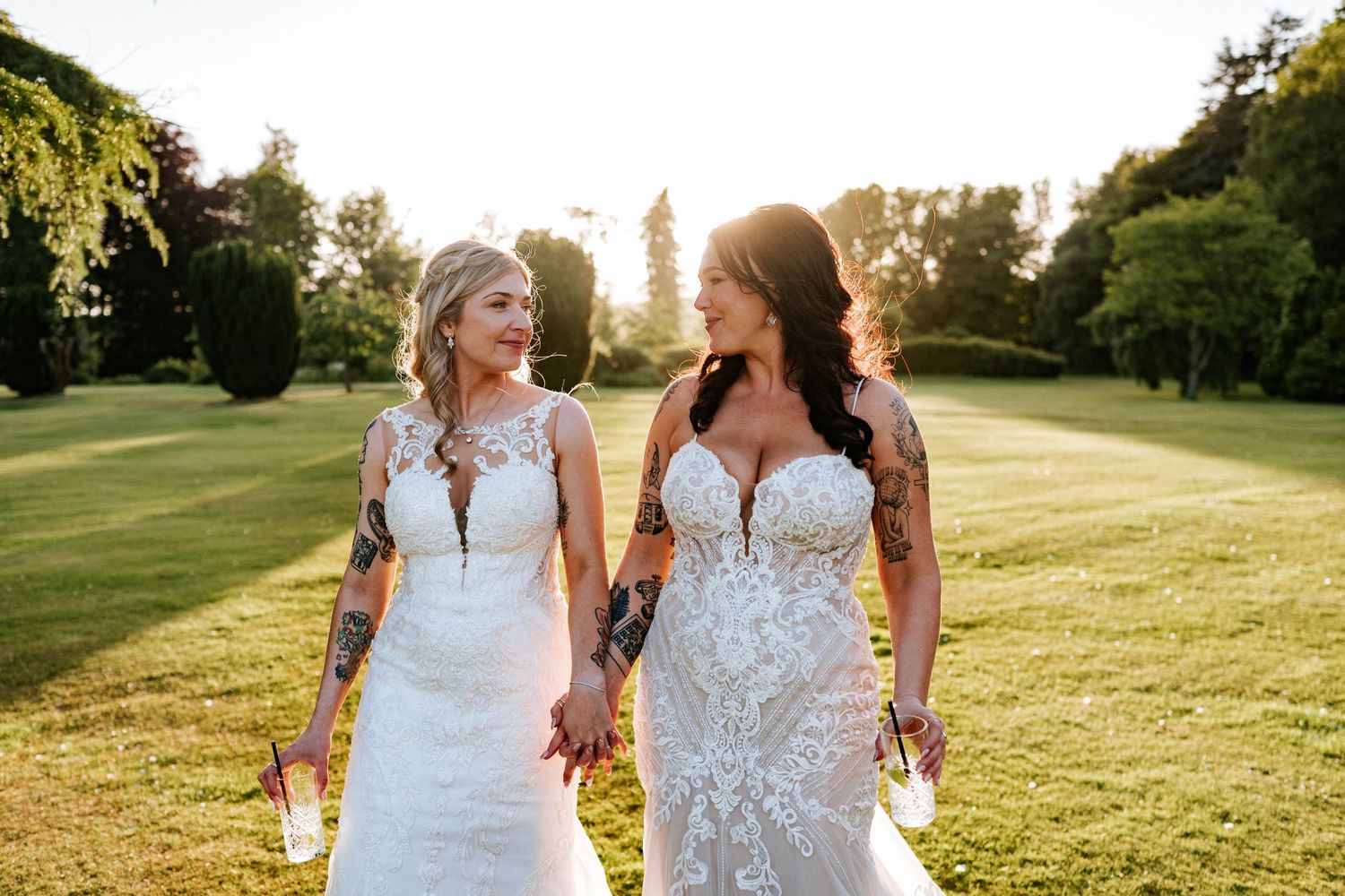 Two people in white dresses walk together through a sunlit grassy field at golden hour.