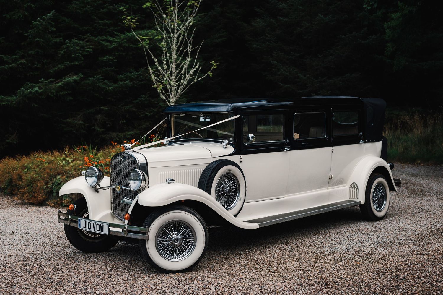 Classic vintage white and black Rolls Royce wedding car parked on gravel path with forest backdrop.