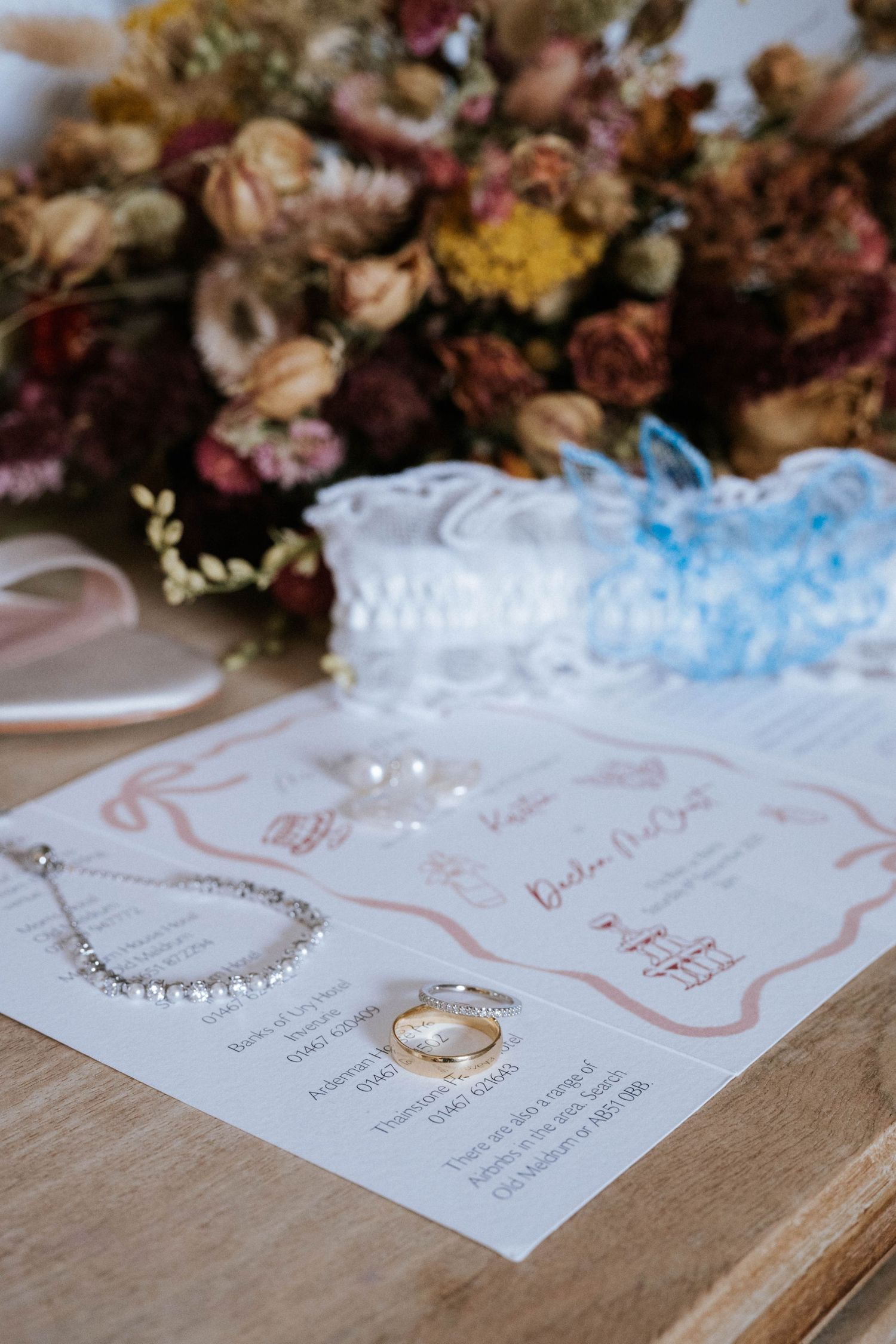Wedding invitation with rings and jewelry displayed alongside dried floral arrangement.