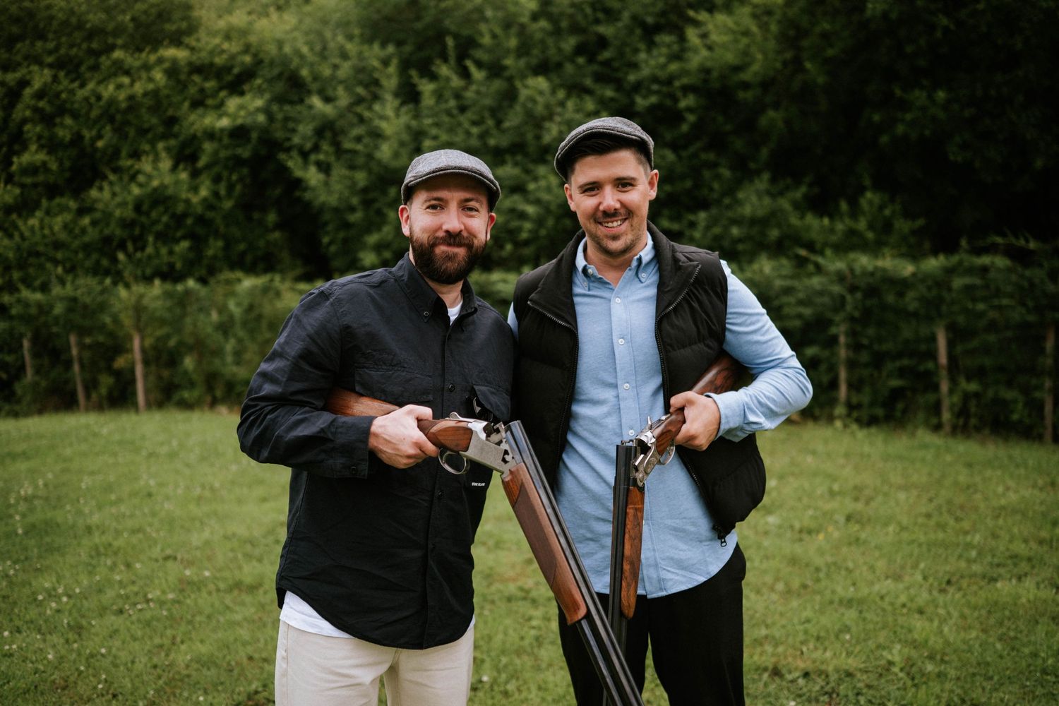 Groomsmen in dark jackets and flat caps pose with hunting rifles in a green field.