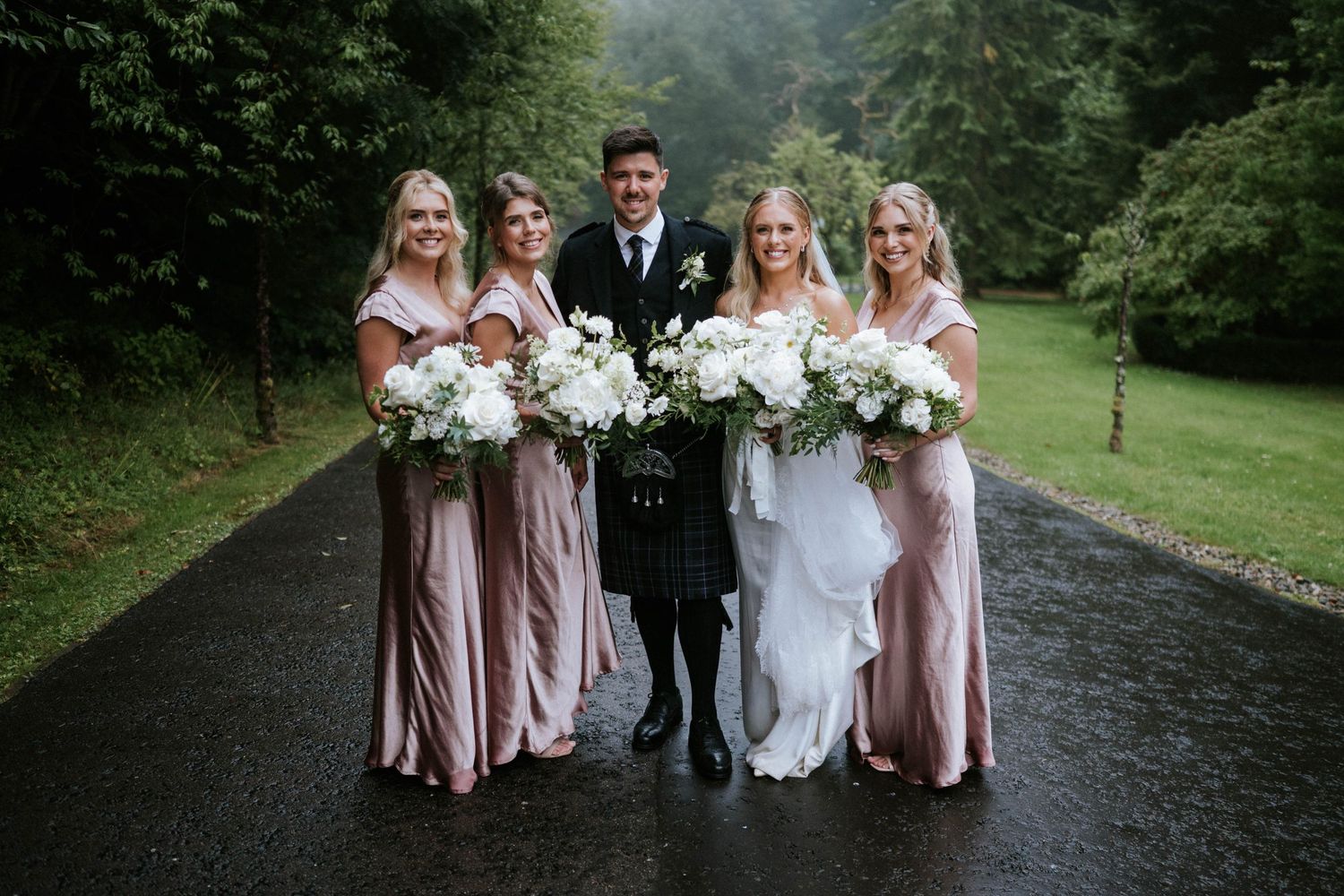 Wedding party members in pink dresses and formal attire pose on a stone path outdoors.