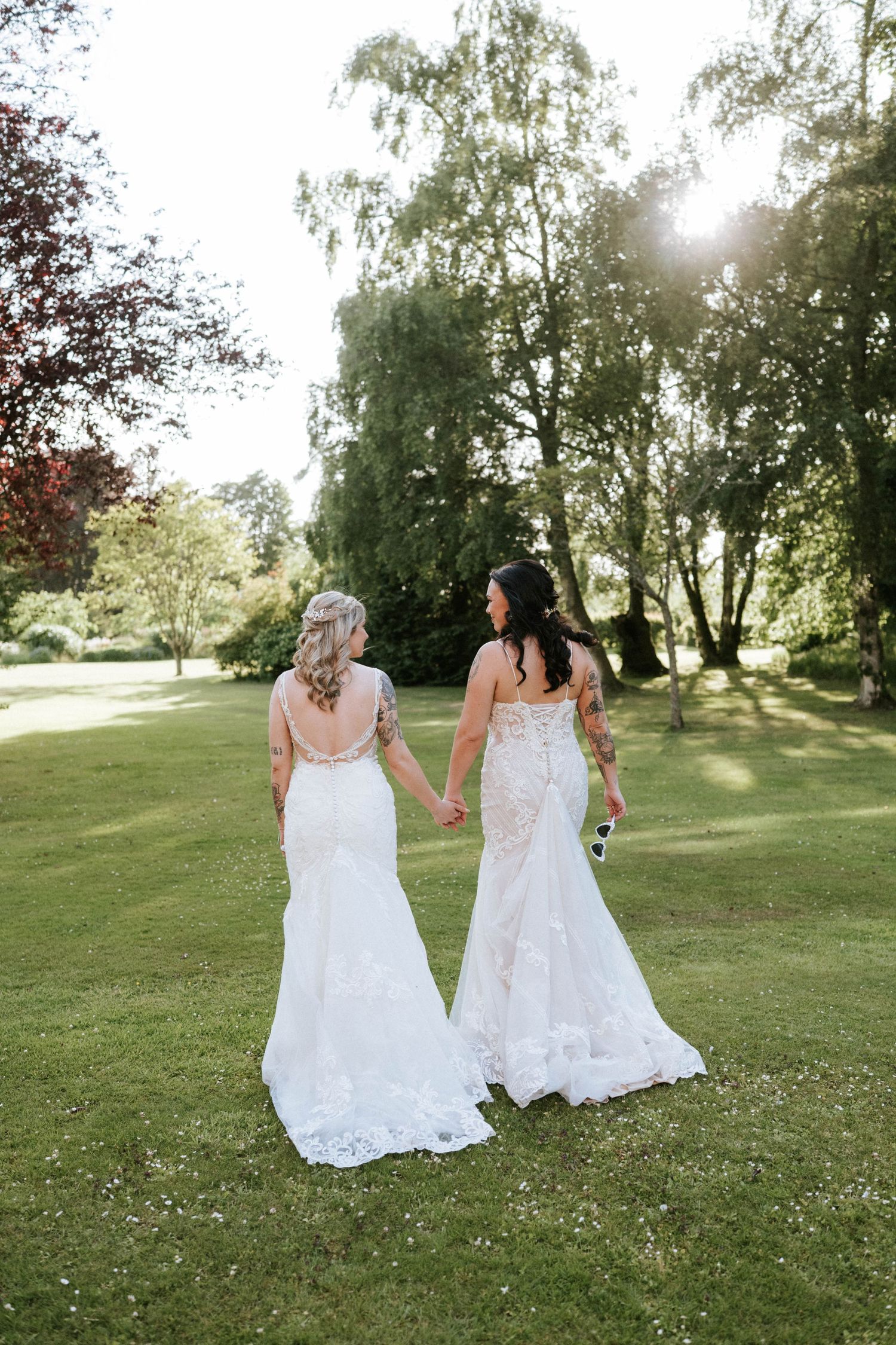 Aberdeen Wedding Photography. Two brides in white lace dresses walking hand in hand through a sunlit garden.