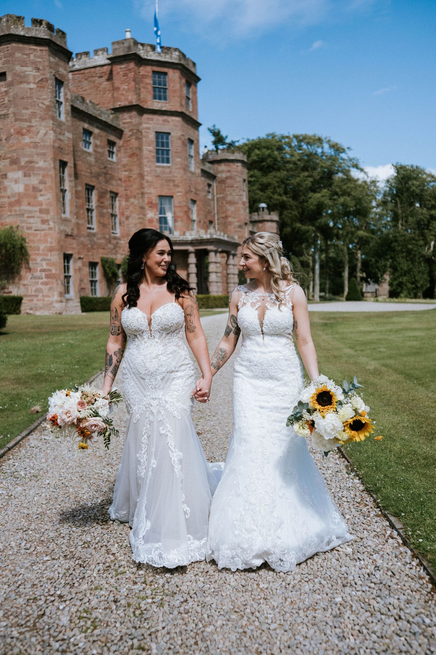 Two brides in elegant white lace gowns walking hand in hand outside a historic castle.