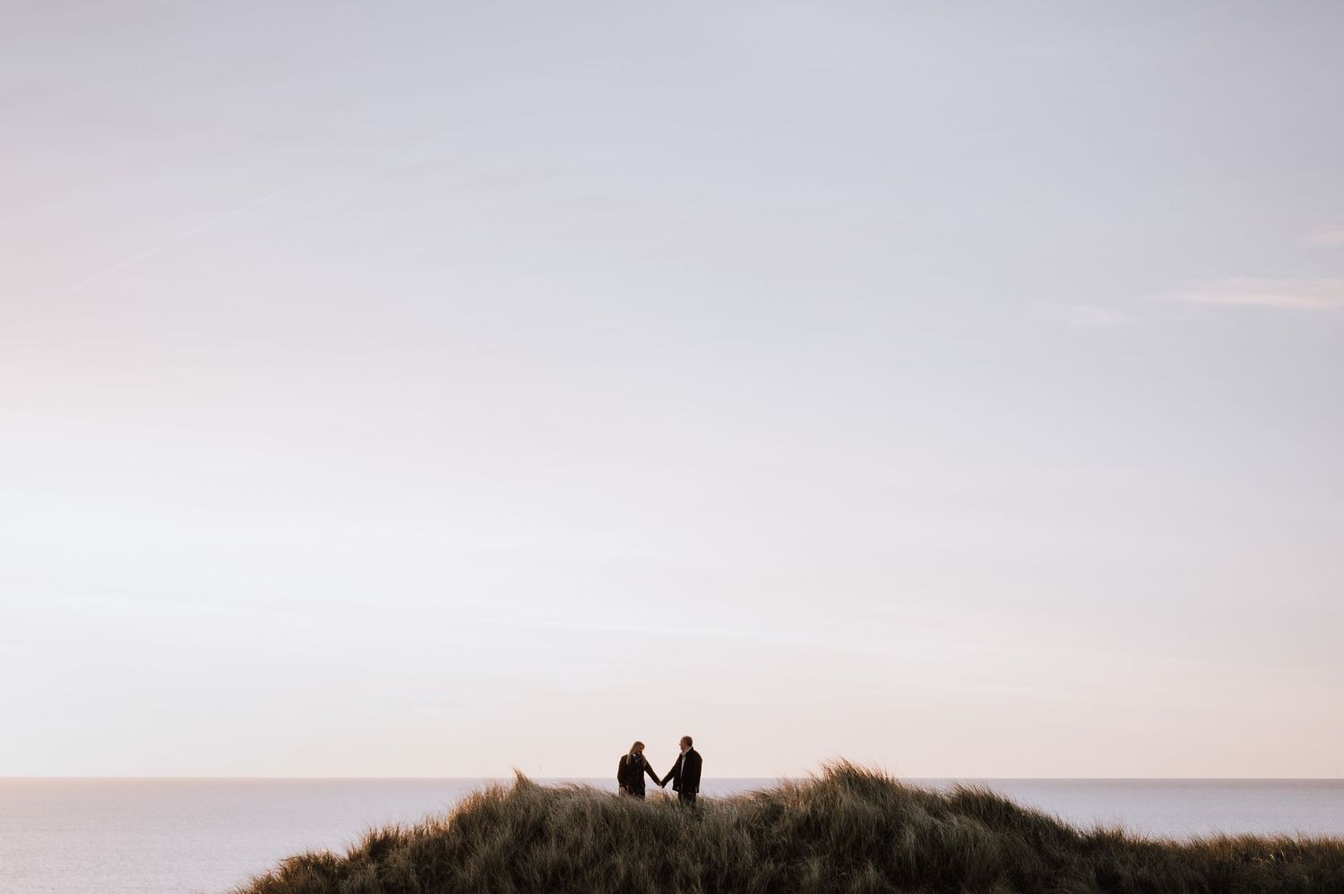 A scenic mountain landscape with silhouettes of two hikers standing on the summit against a cloudy sky at sunset.