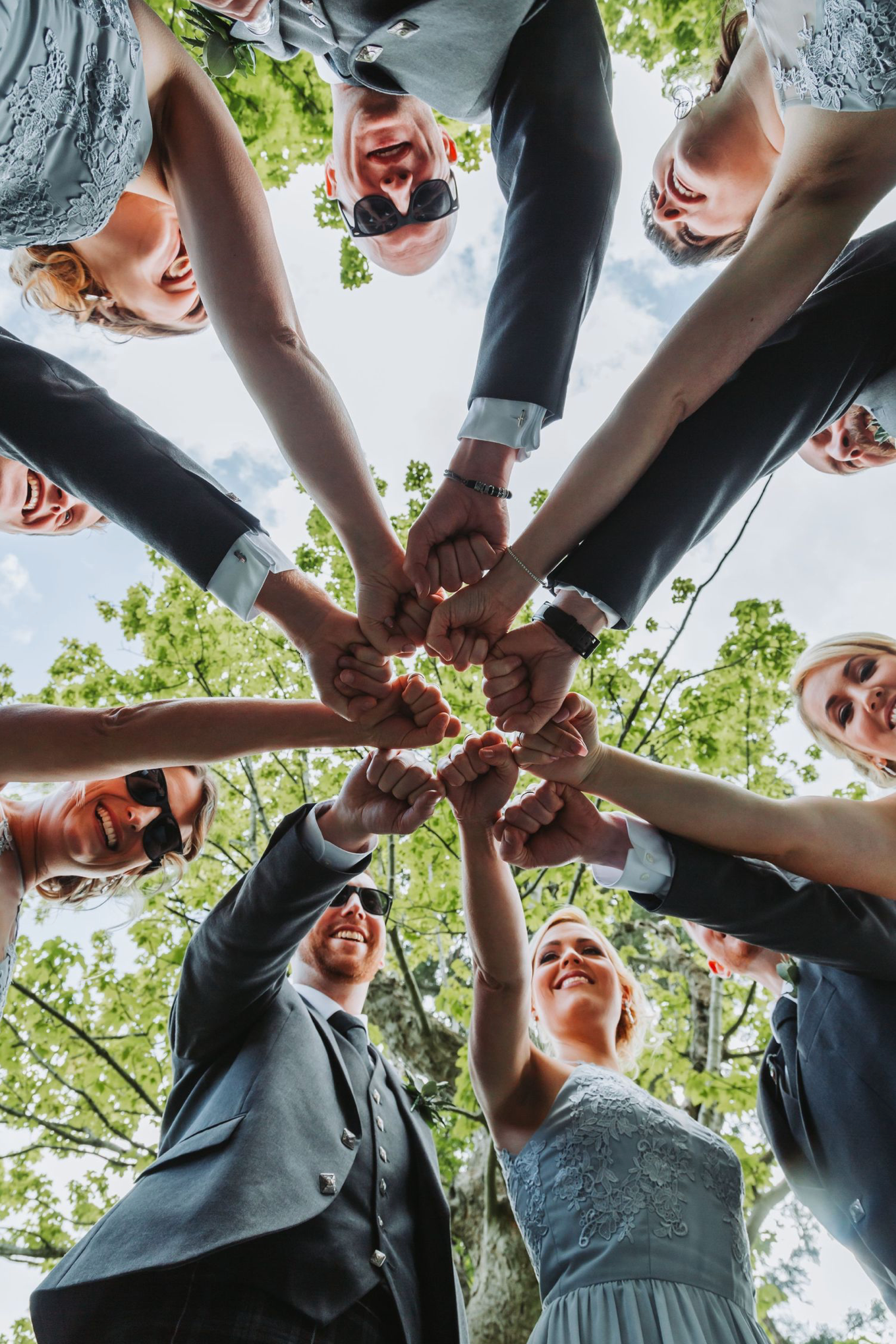 A group of people in formal attire join hands in a circle formation viewed from below against a green leafy backdrop.