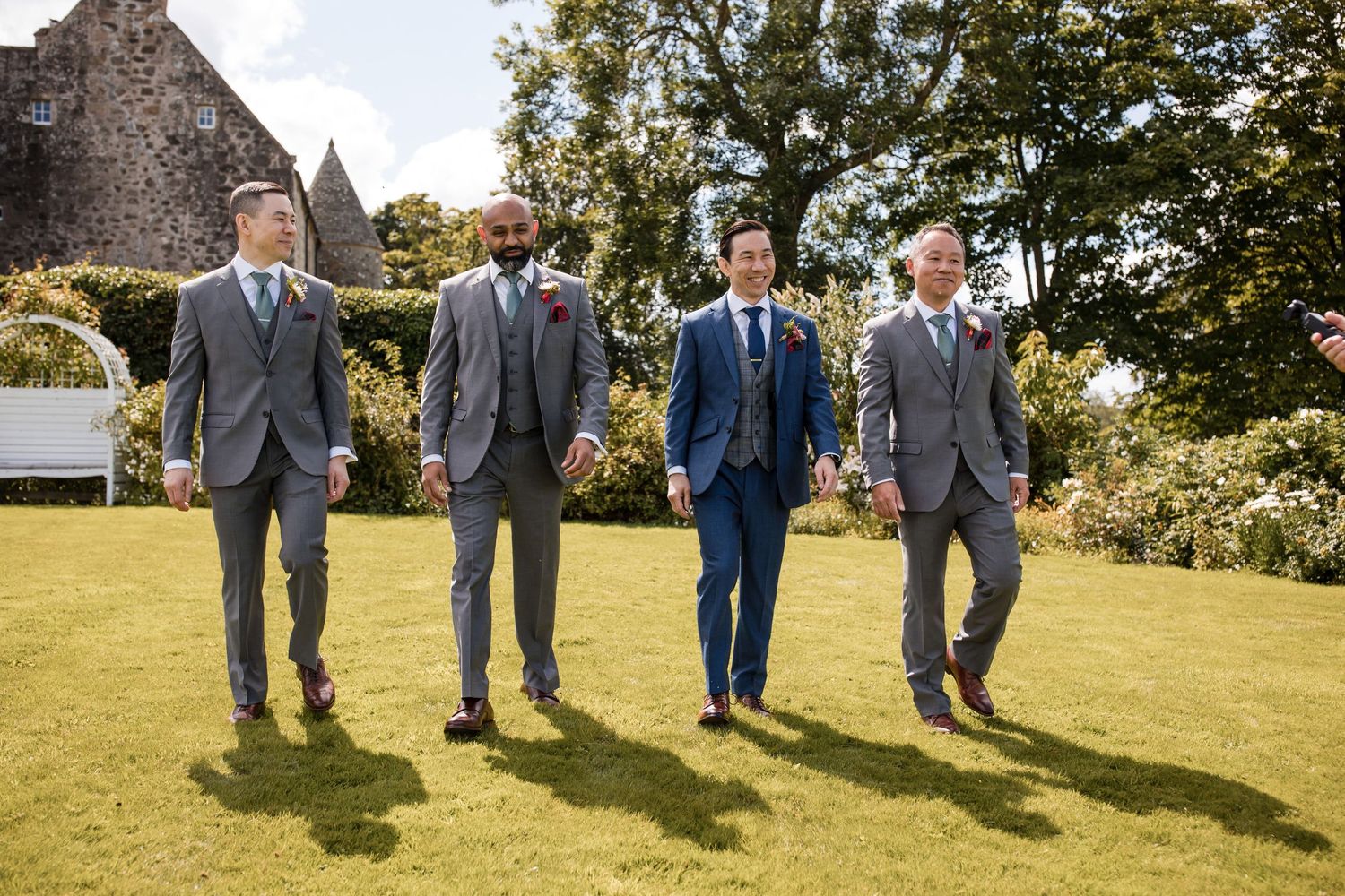 Four groomsmen in coordinated suits walk together on a sunny lawn.