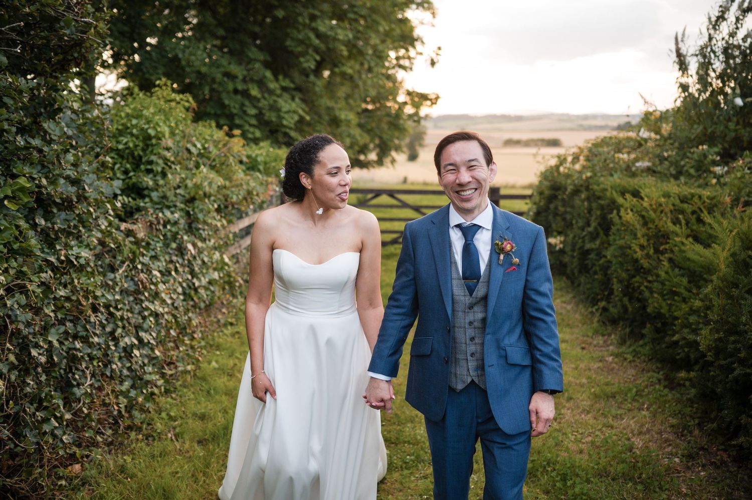 A couple walks together on a countryside path surrounded by greenery and rolling hills in the distance.