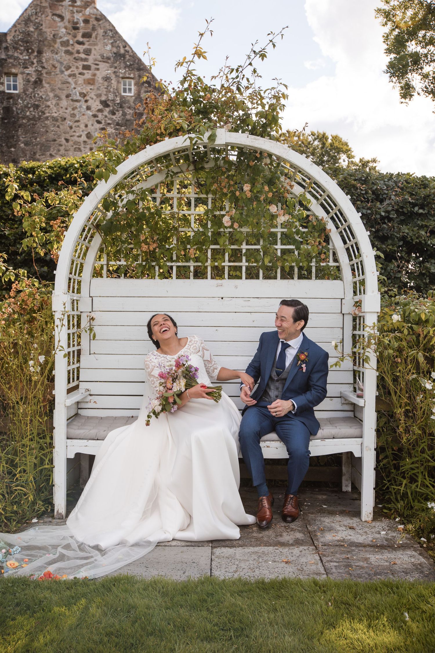 Romantic wedding couple shares sweet moments together on a white garden arbor bench against stone wall backdrop.