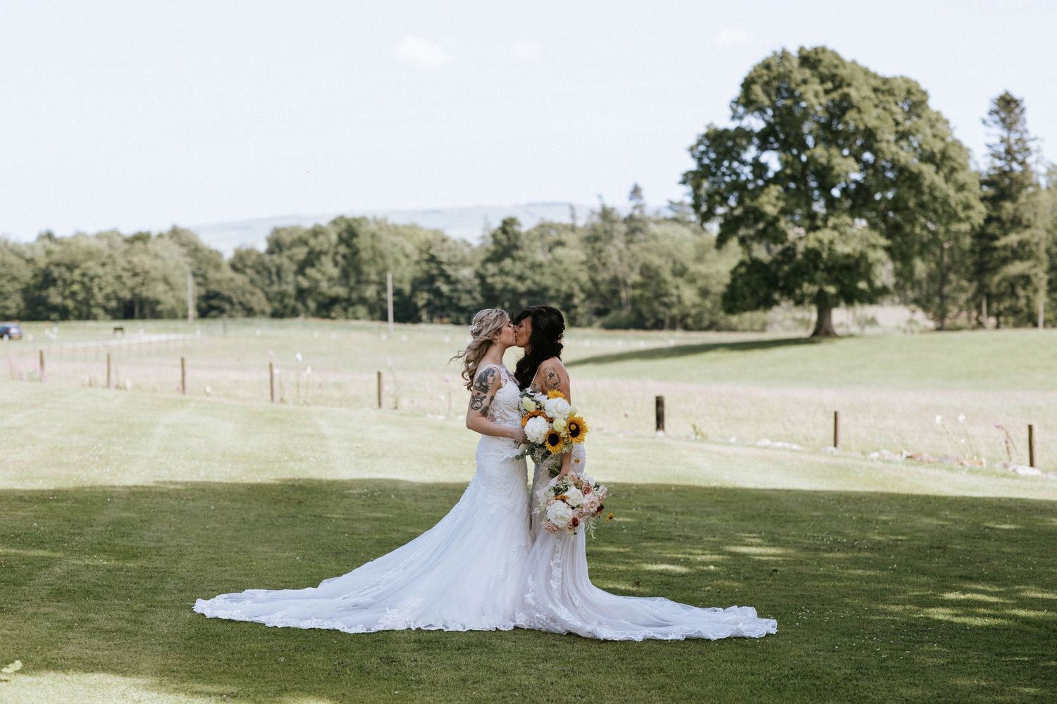 Two brides in flowing white gowns embracing on a sunny lawn with bouquets.