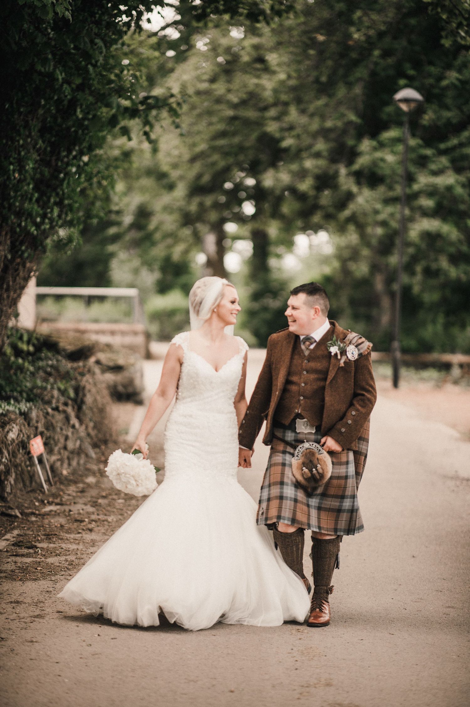 A couple walks hand in hand along a garden path, with the groom in traditional Scottish kilt attire.