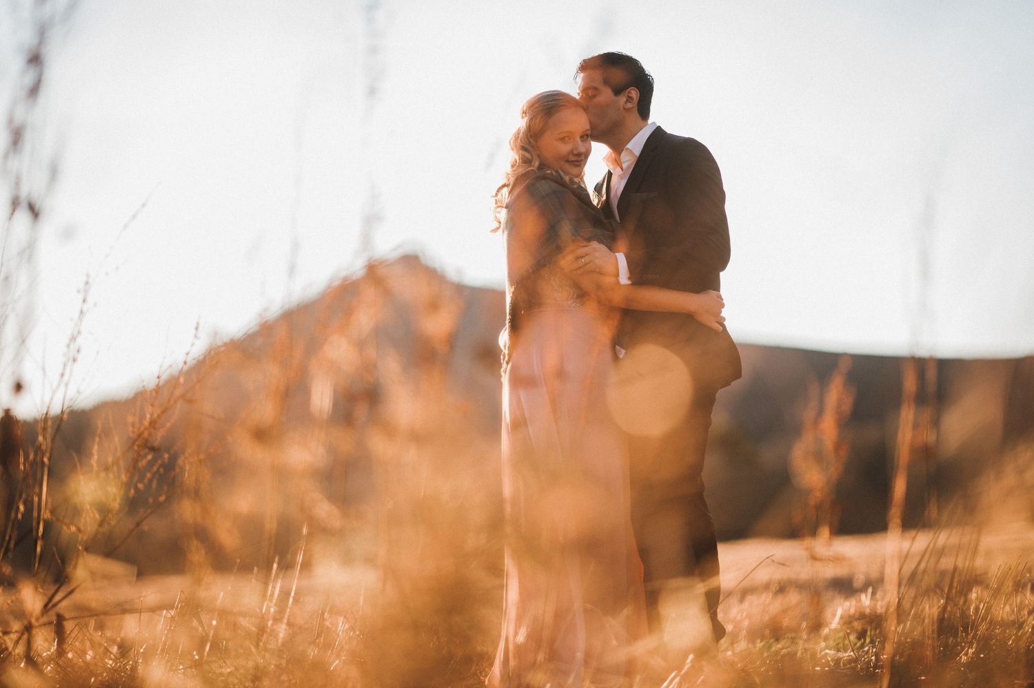 A romantic moment captured in warm sunlight with mountains and tall grass.