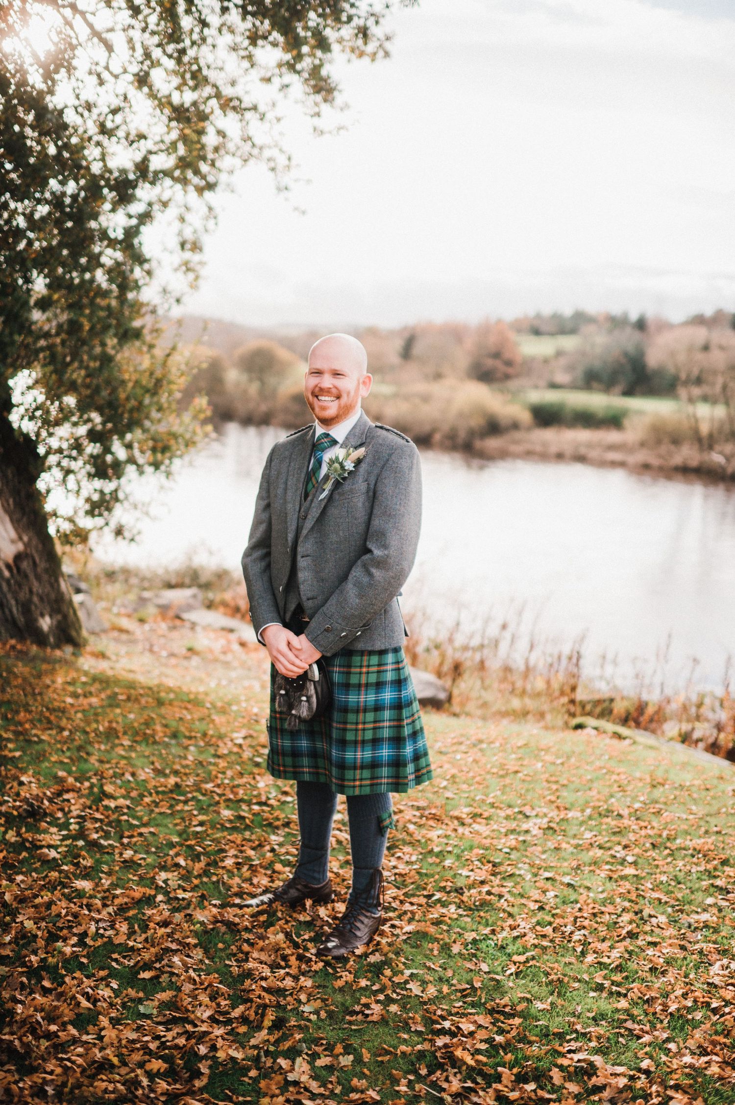 A person in grey formal wear and tartan kilt stands on autumn leaves by a lake.
