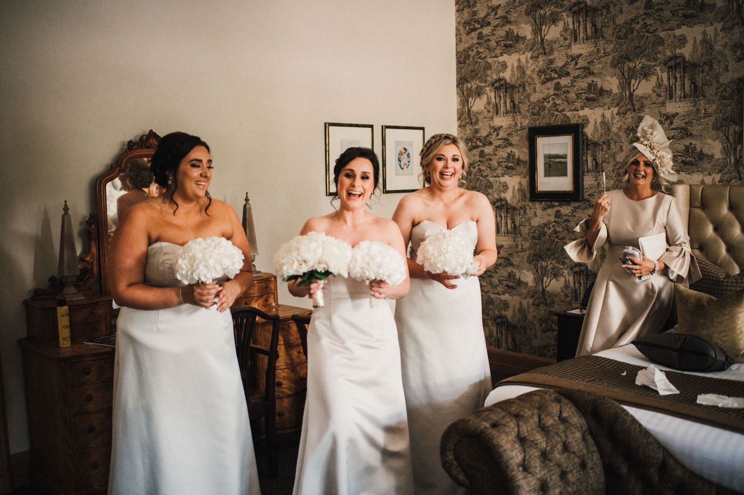 Four bridesmaids in elegant white dresses hold bouquets while standing in an indoor venue with stone walls.
