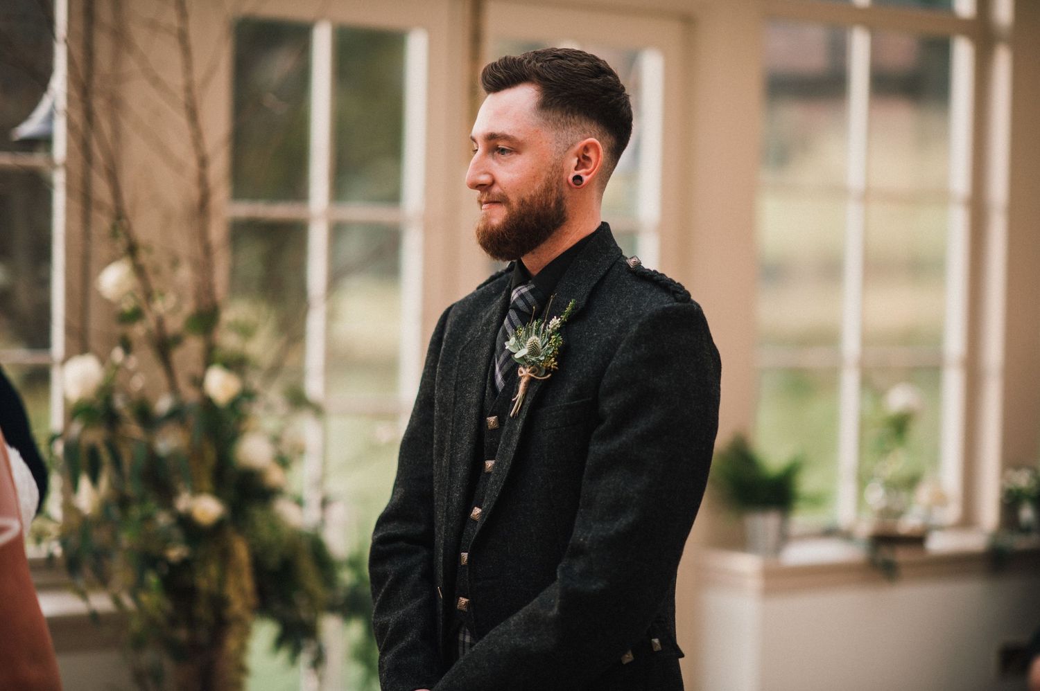 A groom in traditional Scottish attire smiles during an indoor wedding ceremony.