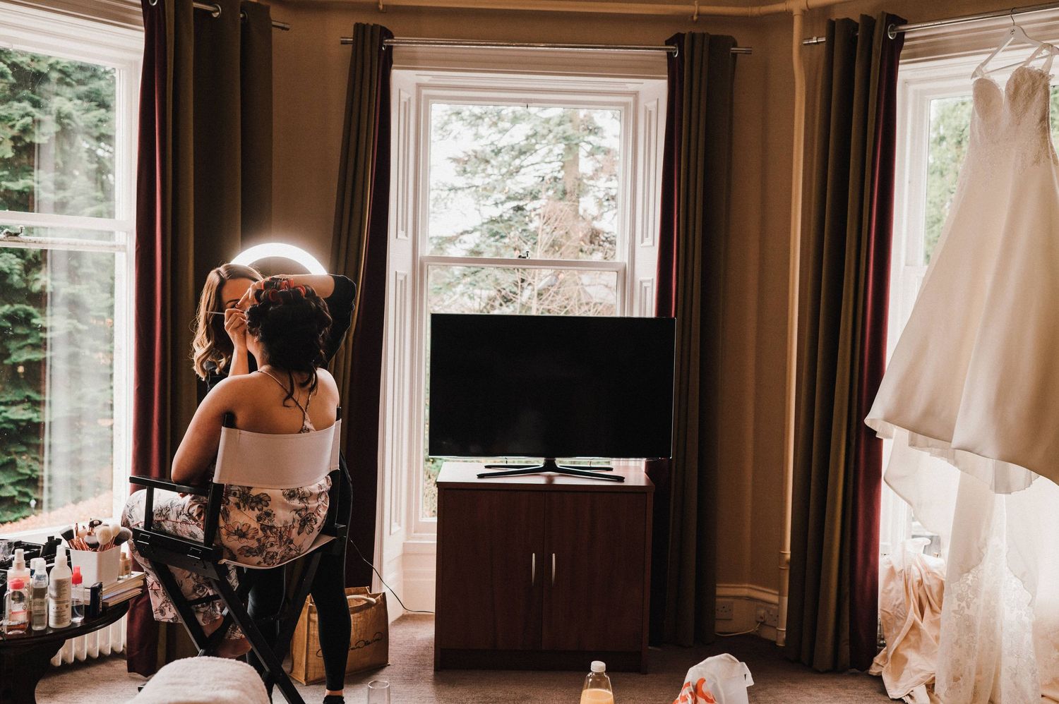 Getting ready in a warm-lit room with a white wedding dress hanging by the window.