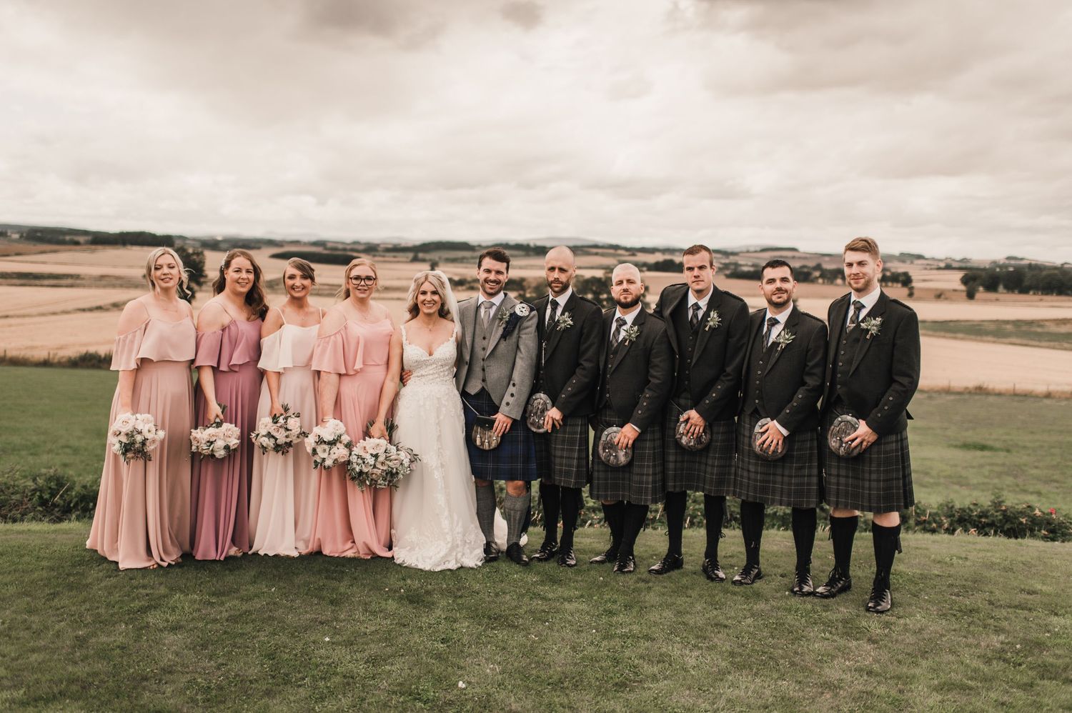 Wedding party in traditional Scottish attire with bridesmaids in pink dresses and groomsmen in kilts pose outdoors.