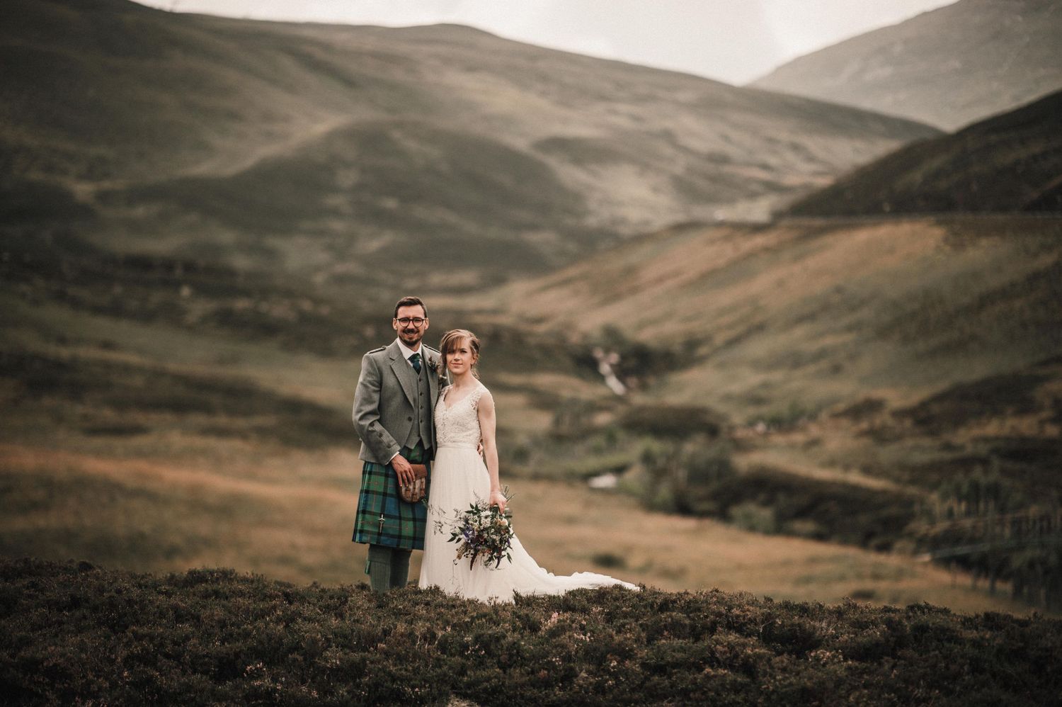 A couple in traditional Scottish attire poses against dramatic rolling hills in the Scottish Highlands.