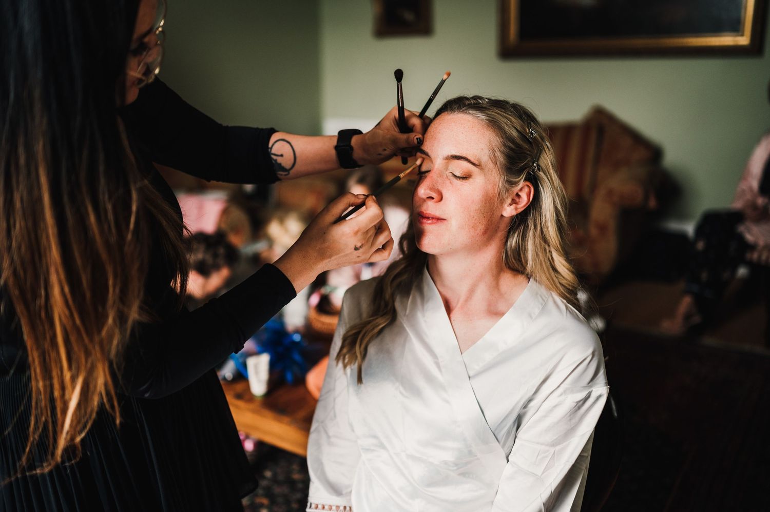 A makeup artist applies cosmetics to a client wearing a white robe during wedding preparations.