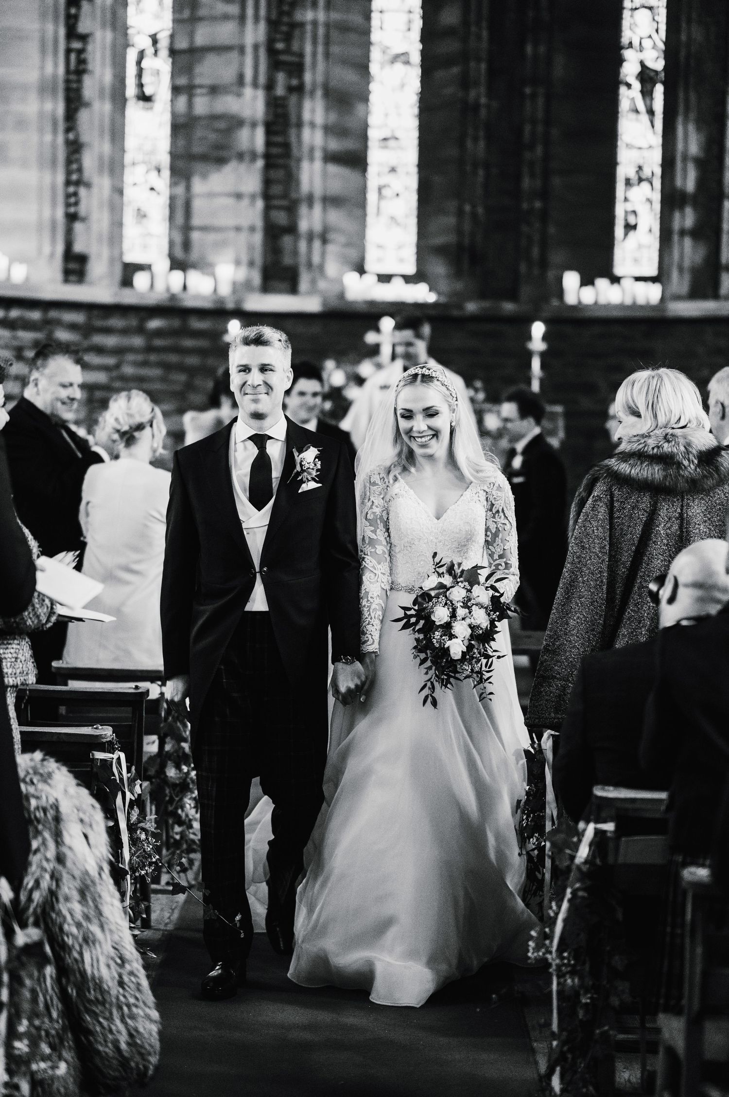 A couple walks down the church aisle in formal wedding attire surrounded by guests and stained glass windows.
