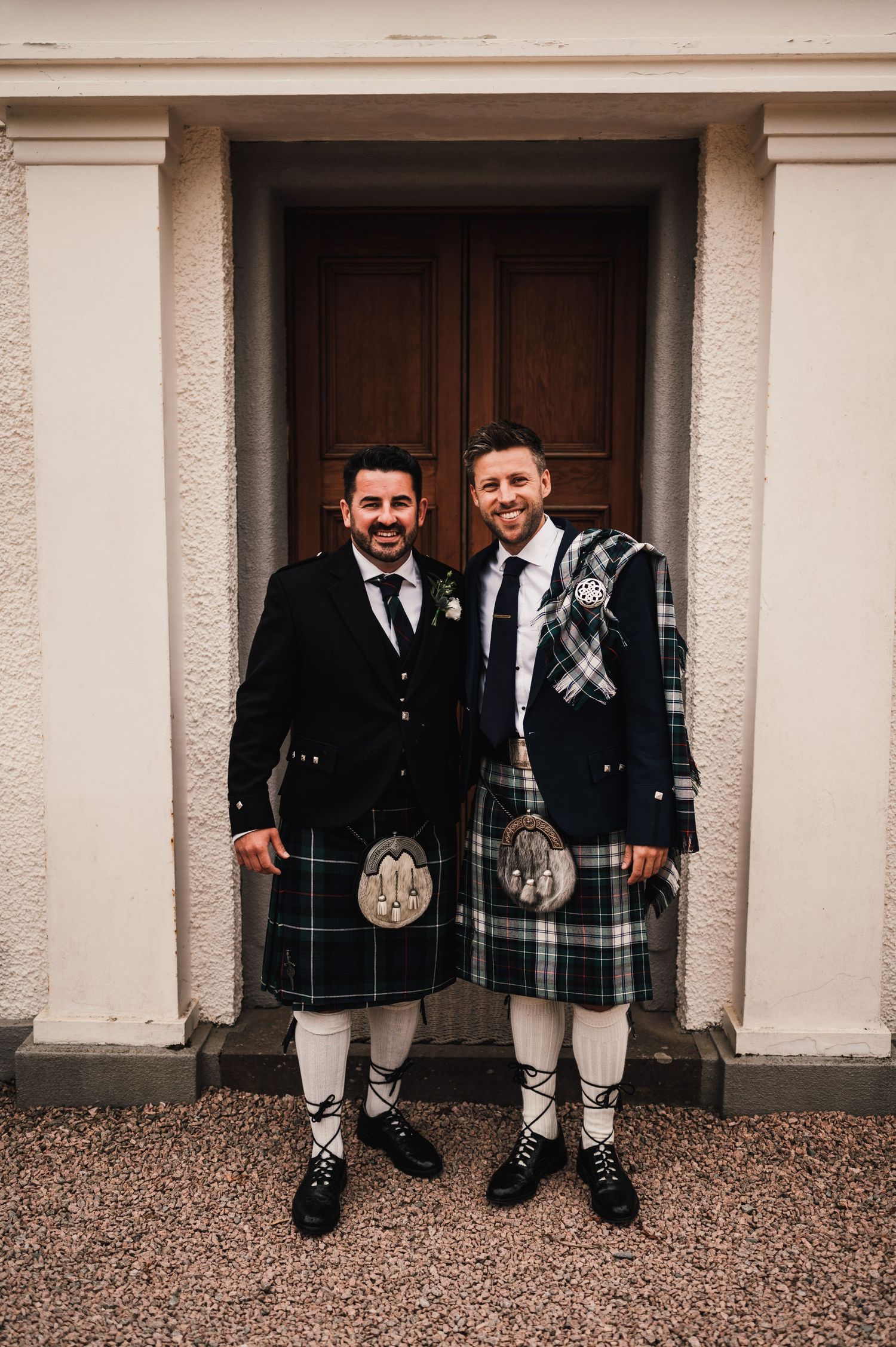 Two men in traditional Scottish kilts and formal wear pose in front of wooden doorway.