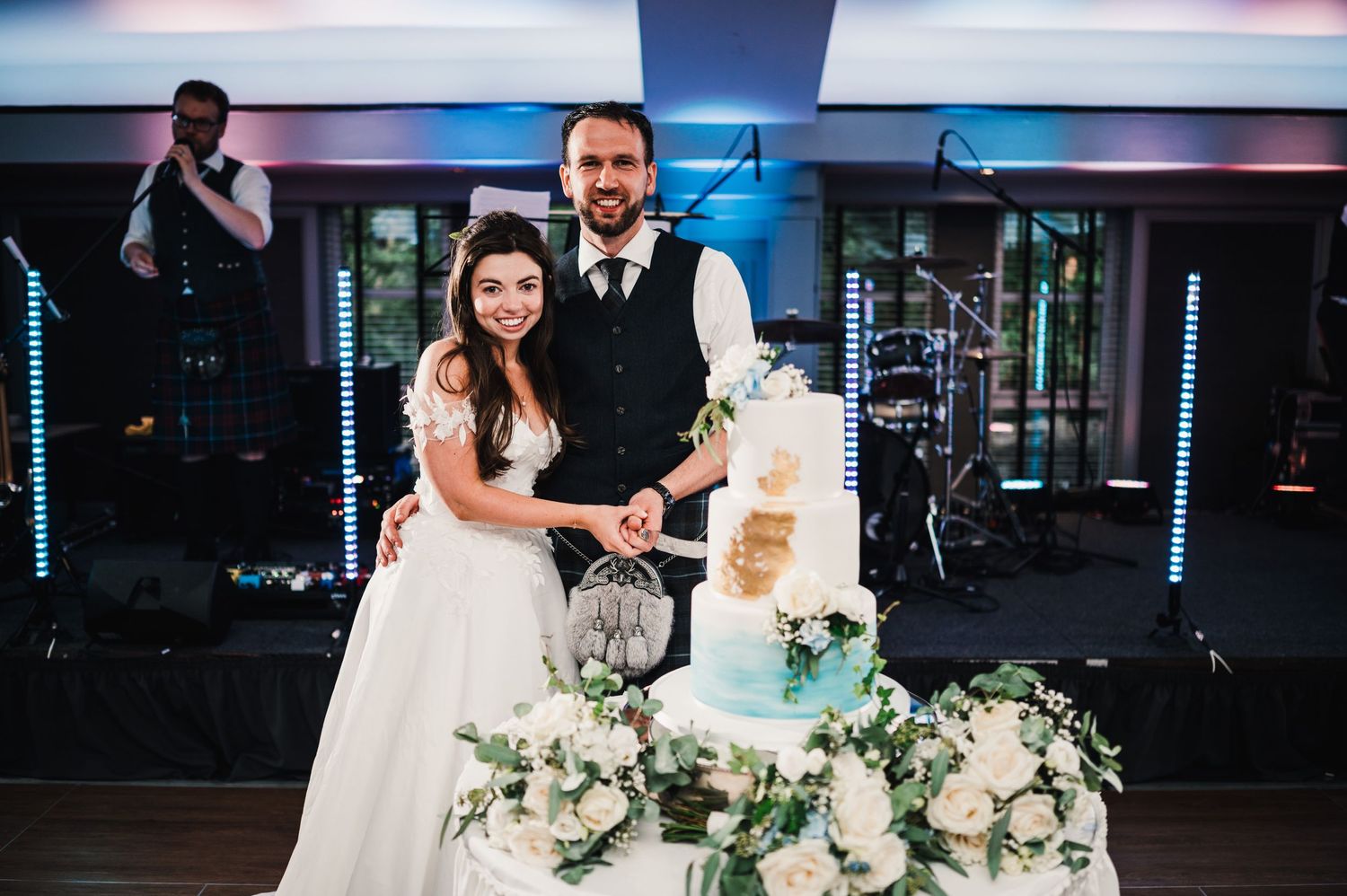 Wedding couple cuts elegant tiered cake decorated with white flowers at reception venue.