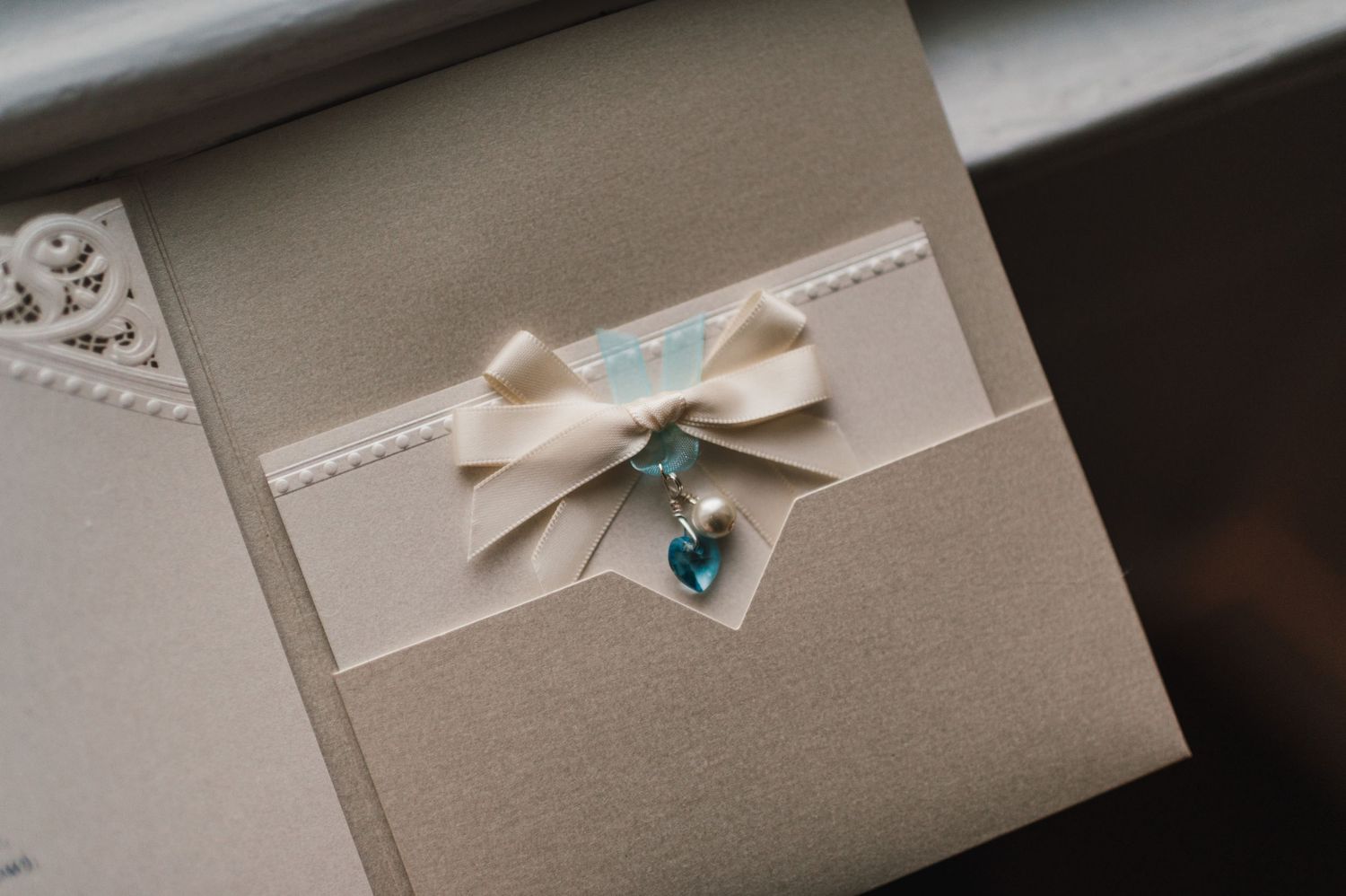 An elegant wedding invitation with cream ribbon bow and blue heart charm on gray paper.
