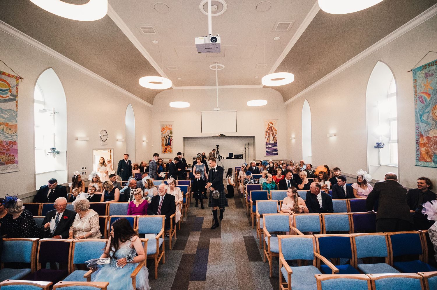 Wide view of a church wedding ceremony with guests seated in blue pews under arched ceilings.