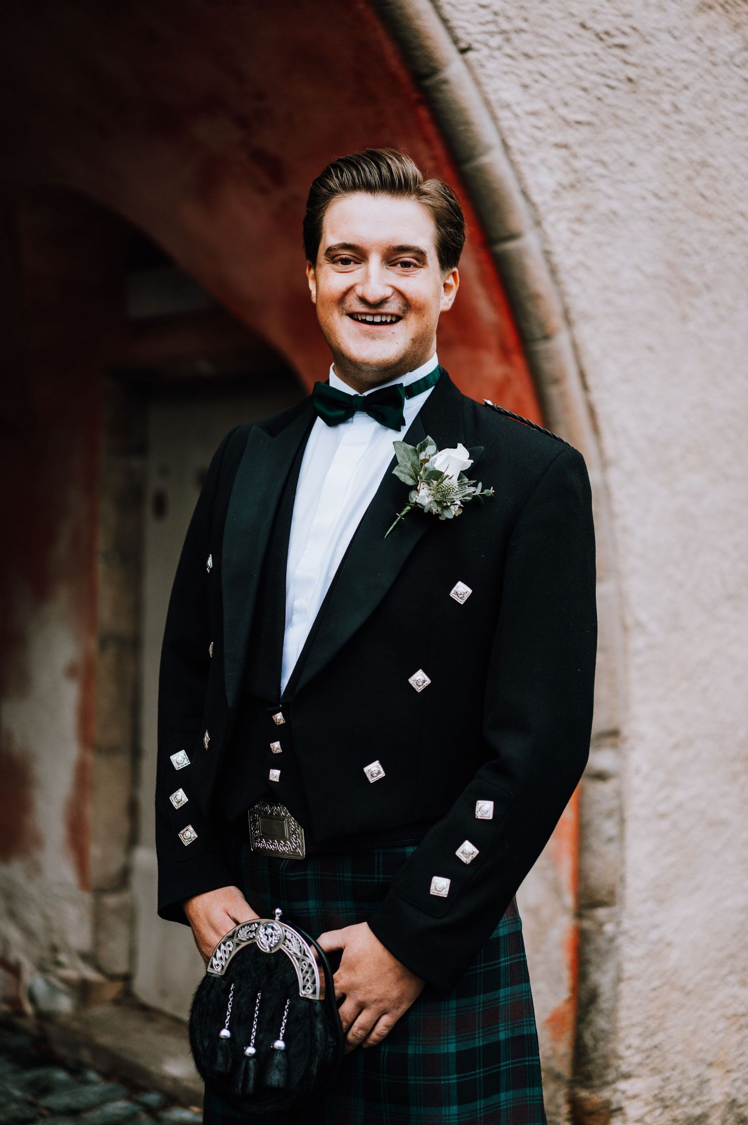 A groom in traditional Scottish formal wear including kilt and bow tie stands smiling under an archway.