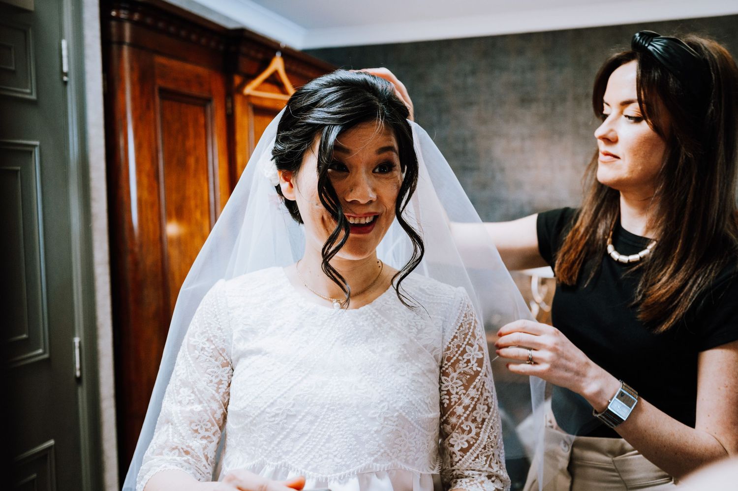 A joyful bride in a white lace wedding dress and veil getting ready with assistance.