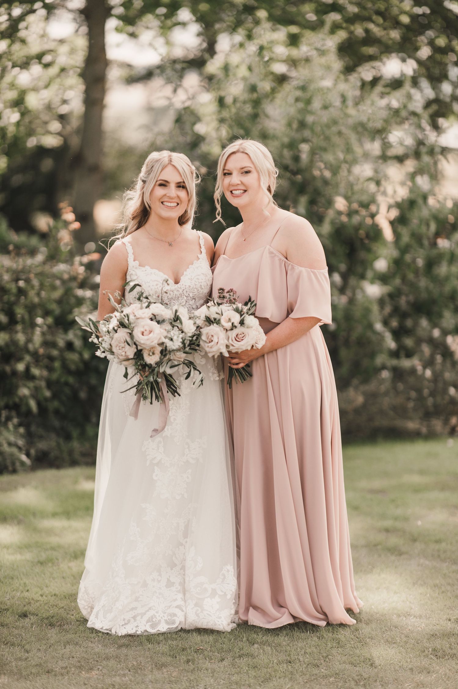 Elegant garden wedding photo with bridesmaid in pink gown and bride in lace dress holding floral bouquets.