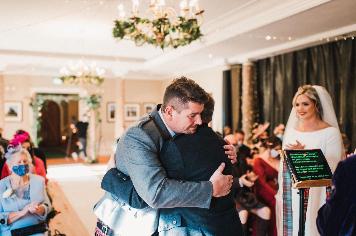 An emotional wedding ceremony moment in an elegantly decorated ballroom with chandeliers and floral decor.