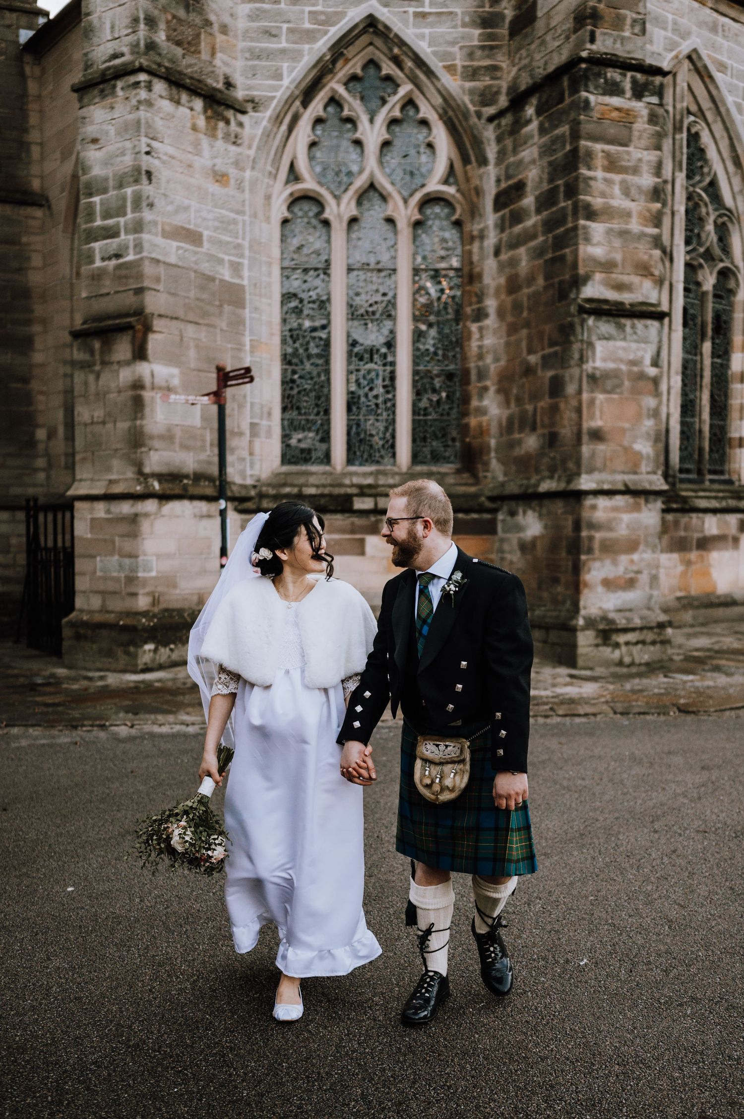 Couple in Scottish wedding attire walks hand in hand by historic church windows.