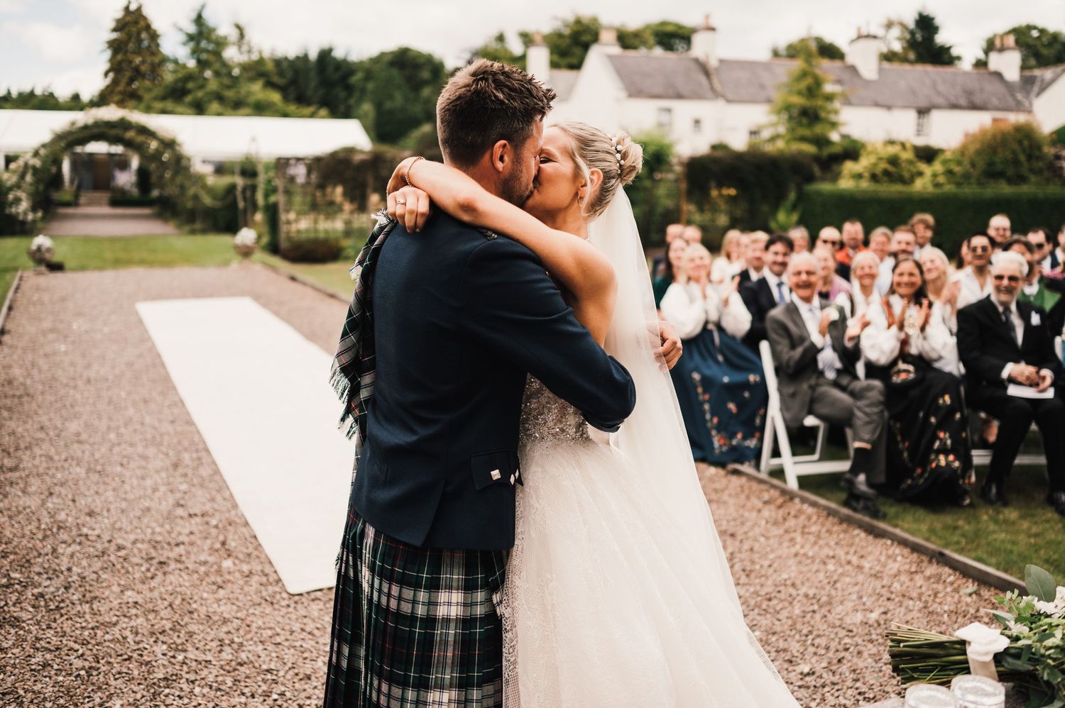 A romantic outdoor wedding ceremony with couple embracing while guests look on from chairs.