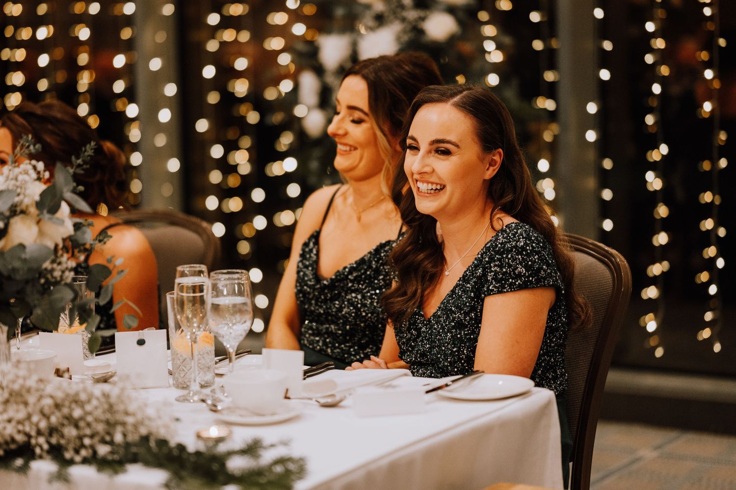 Two guests in sparkly dresses share a joyful moment at a wedding reception with twinkling lights in background.