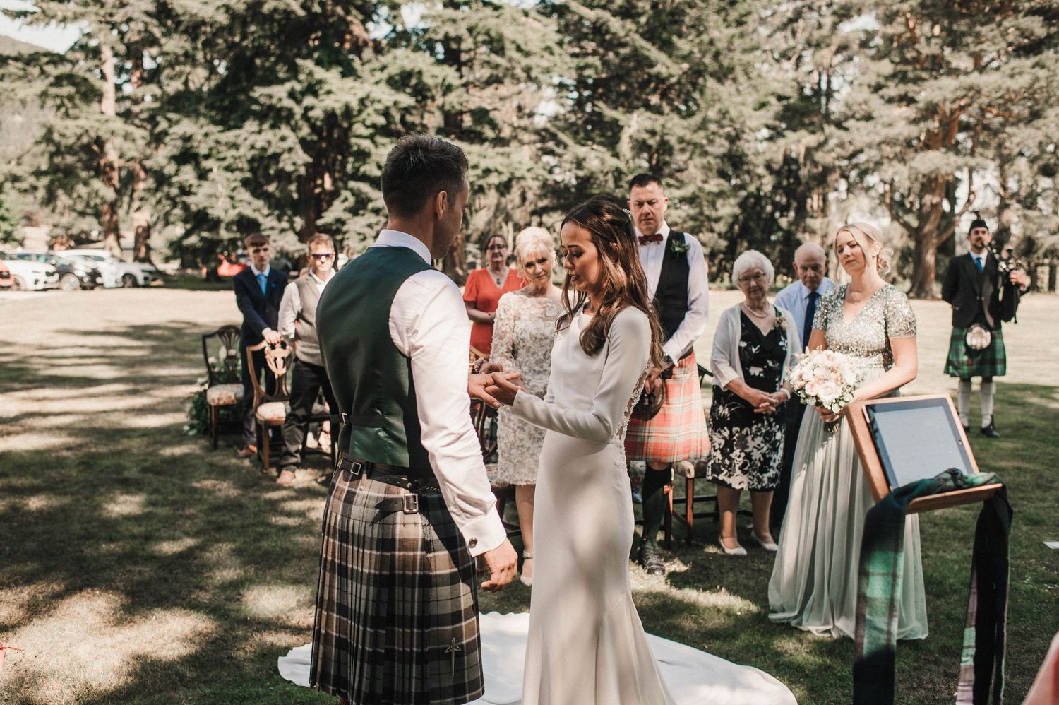An outdoor wedding ceremony takes place in a sunlit forest setting with guests seated nearby.