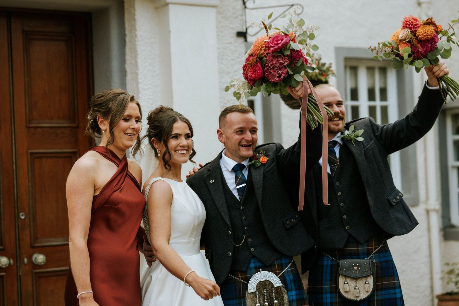 Elegant wedding party gathered by white stone building entrance with burgundy floral accents.