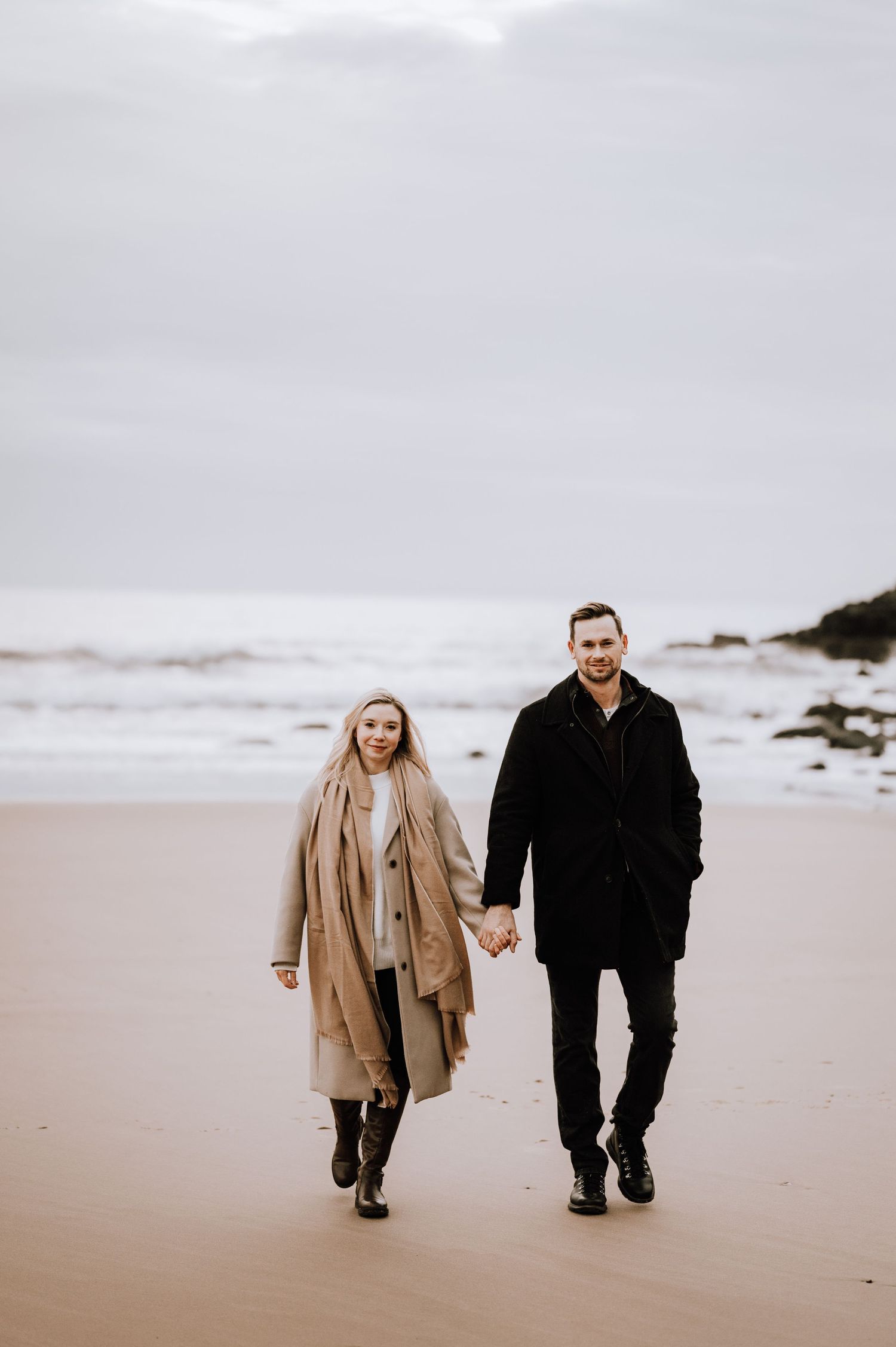 Couple taking a romantic stroll along a sandy beach on an overcast day.