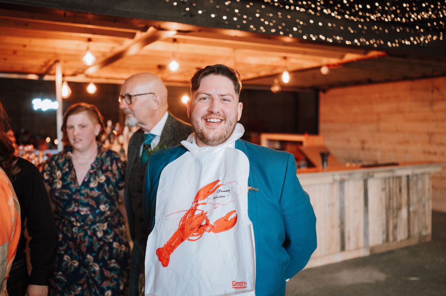 A guest in a blue blazer and lobster shirt smiles during a reception under string lights.