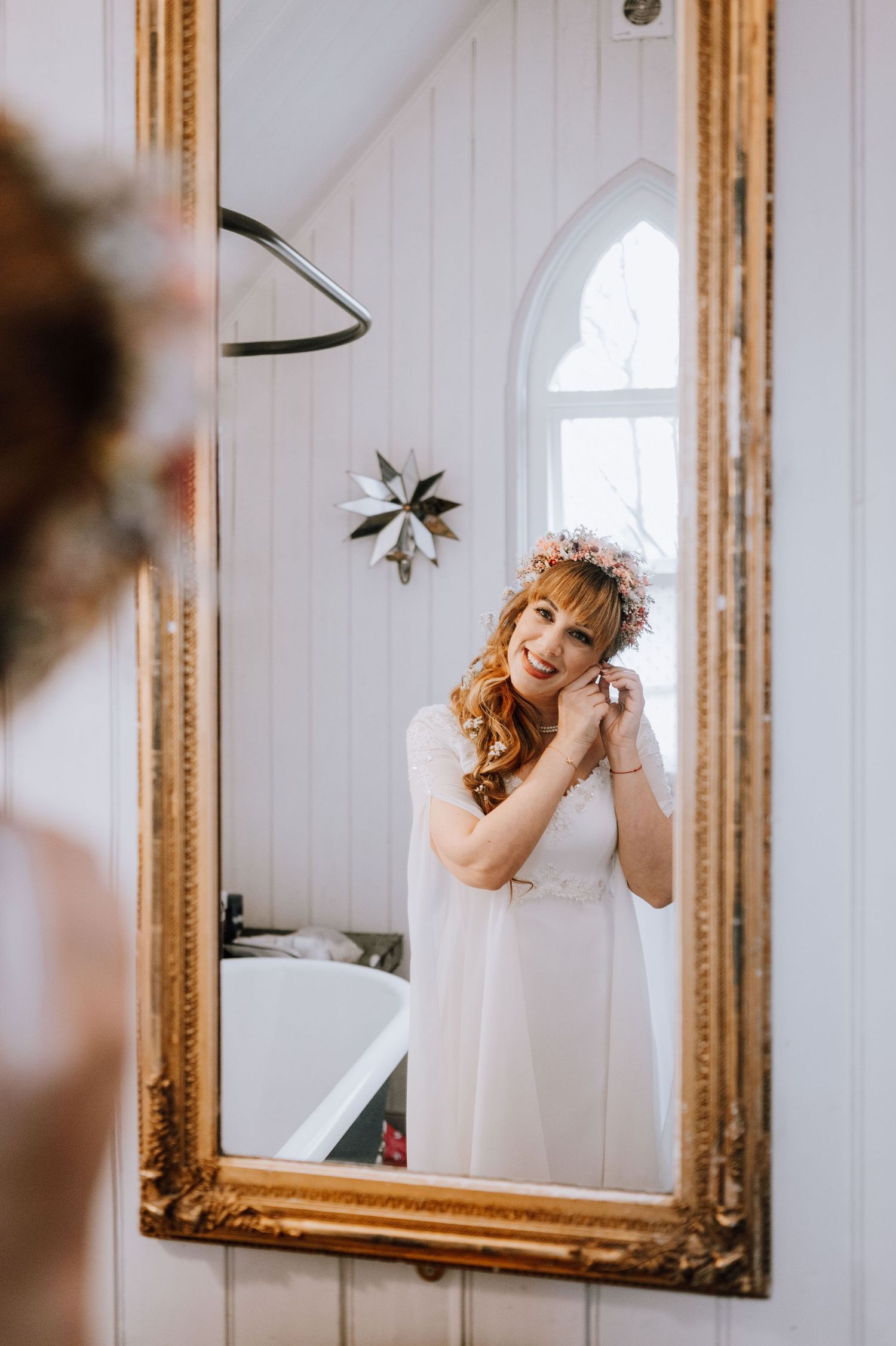 Bride in white dress poses in vintage gold framed mirror with floral crown and flowing curls.