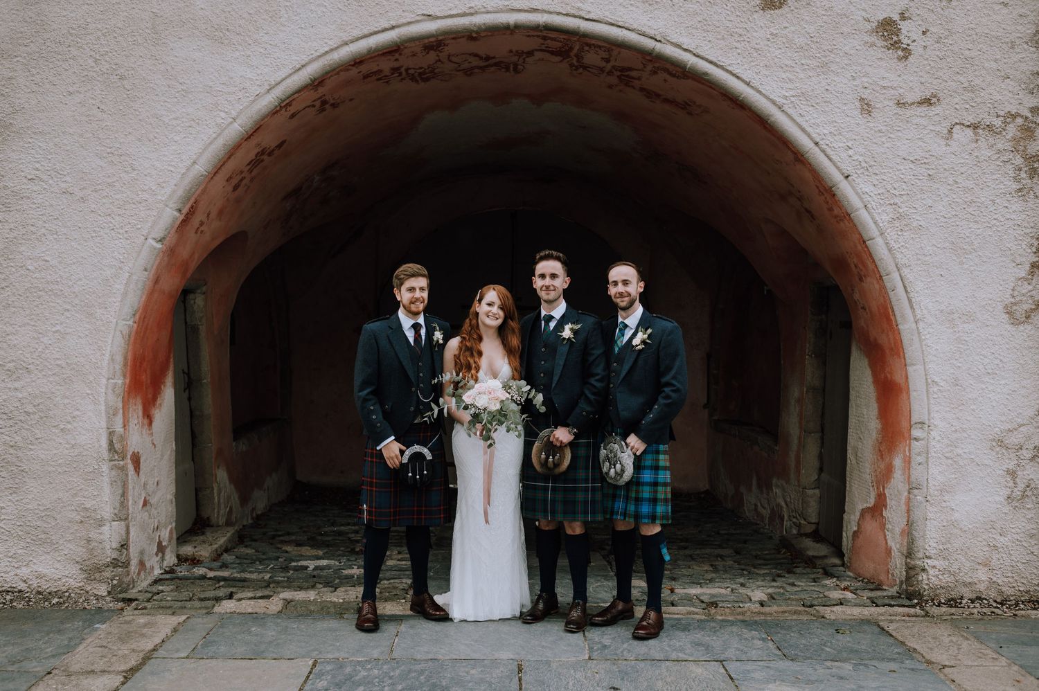 Wedding party in traditional Scottish attire with kilts poses under stone archway.
