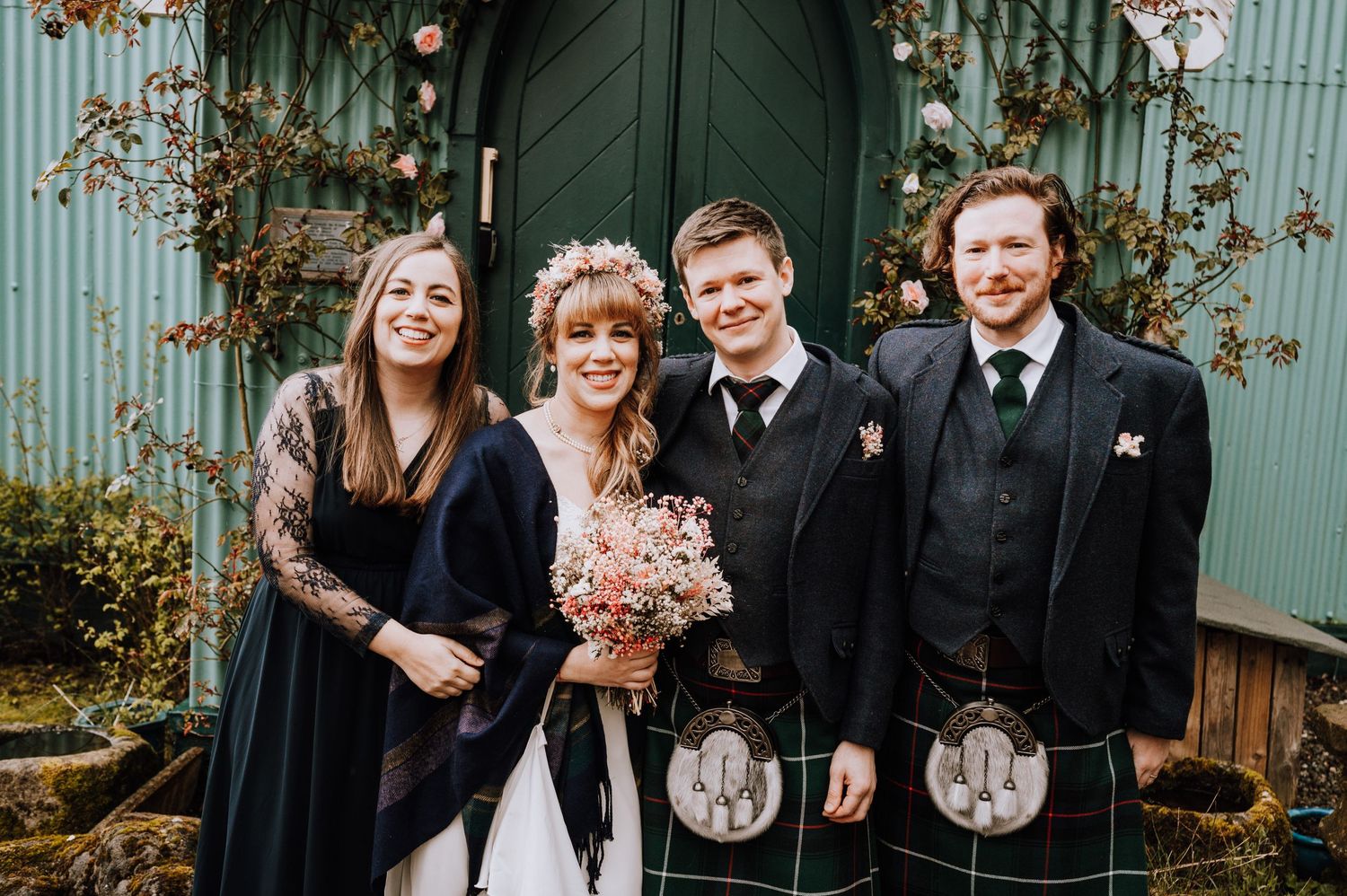 Wedding party wearing kilts and formal attire poses by church entrance.