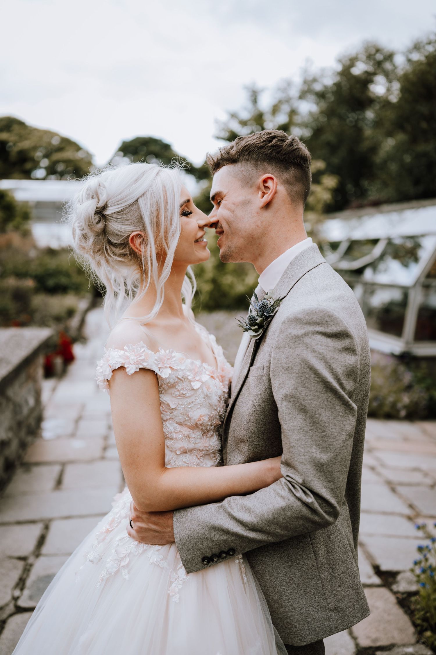 Bride in lace gown and groom in gray suit share a close moment outdoors.