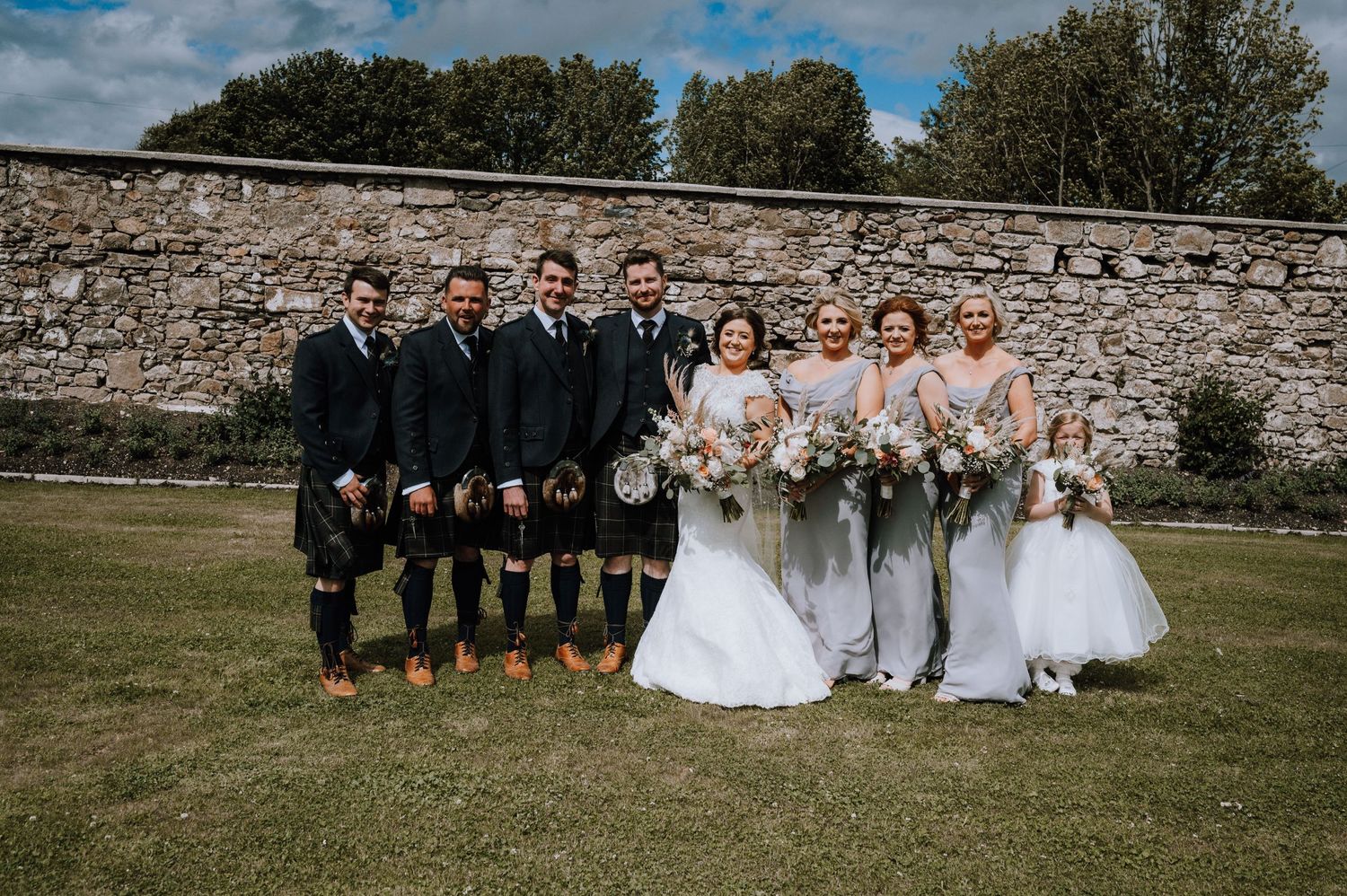 Scottish wedding party in kilts and gowns pose against stone wall outdoors.