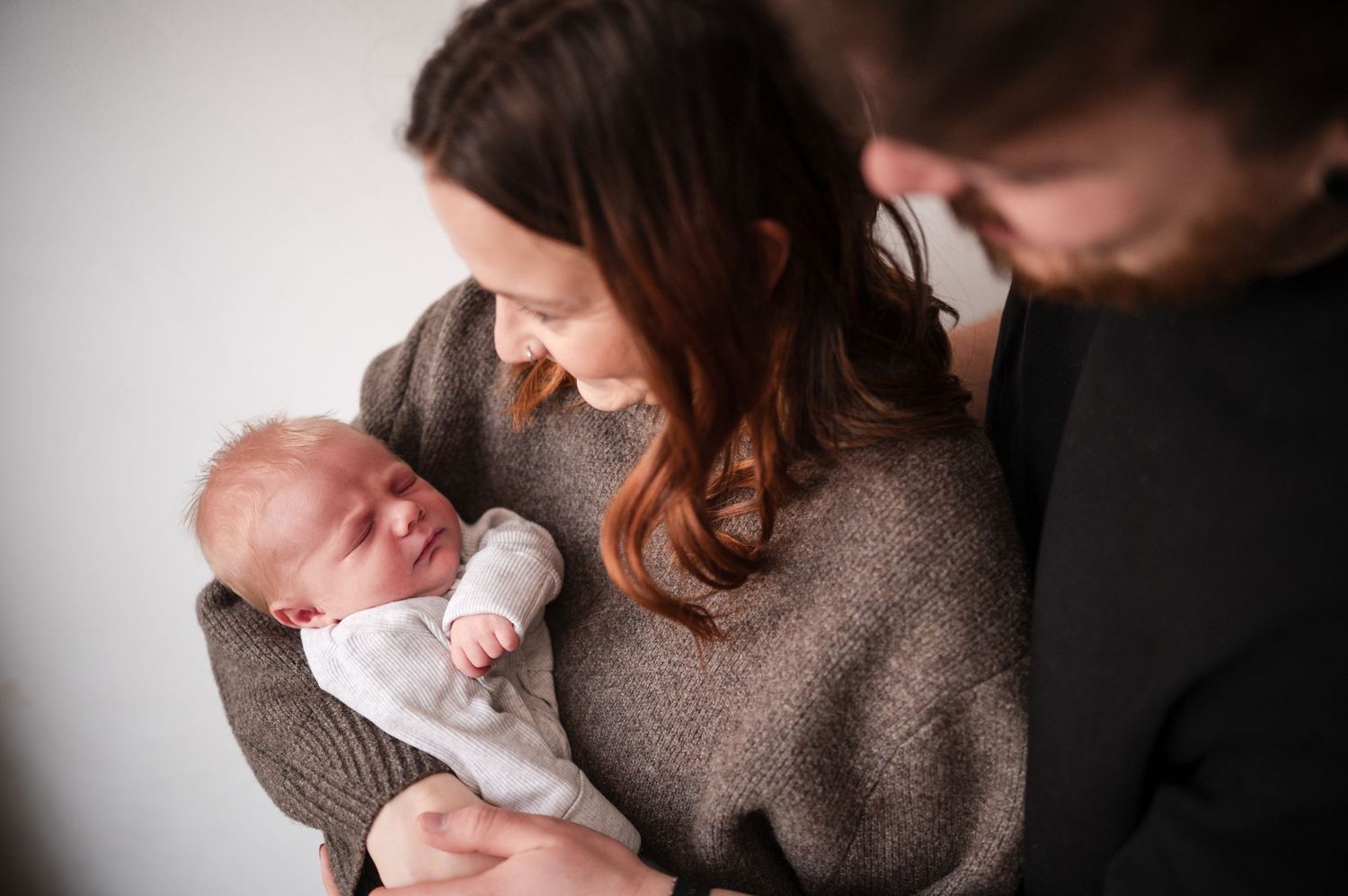 Parents in cozy sweaters cradle and gaze lovingly at their newborn baby against a white wall.
