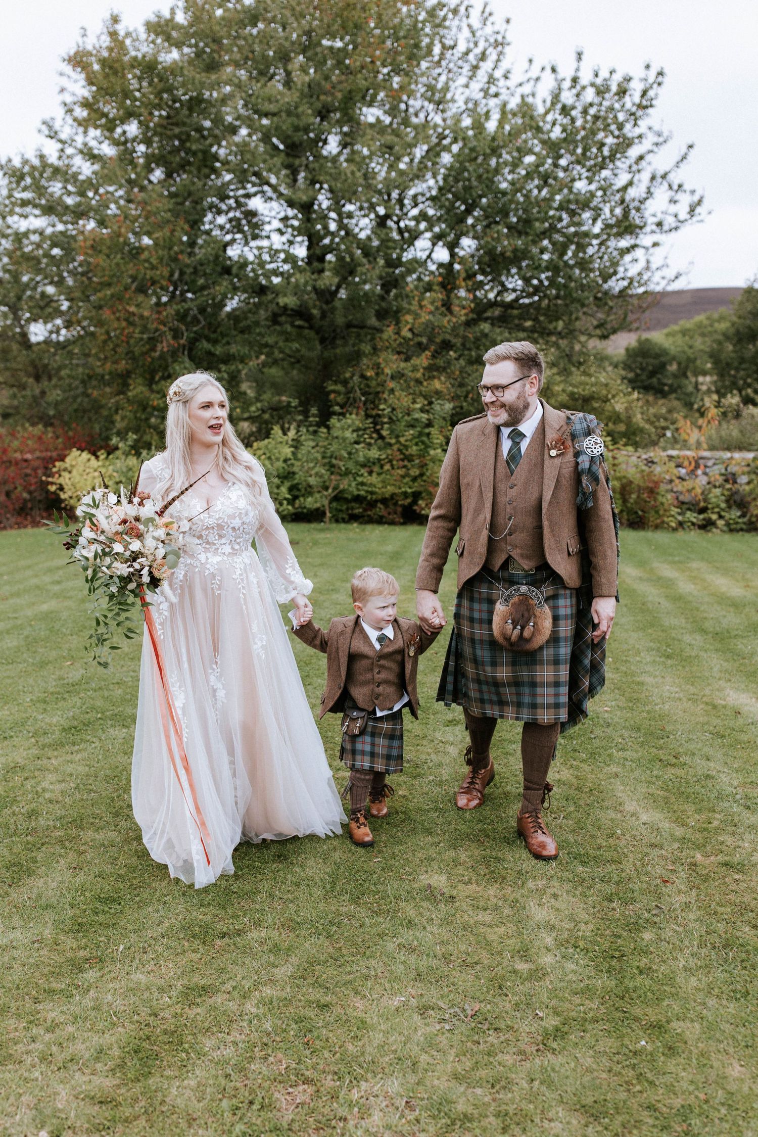 Family walking on lawn with bride in flowing dress and groom in traditional Scottish kilt.