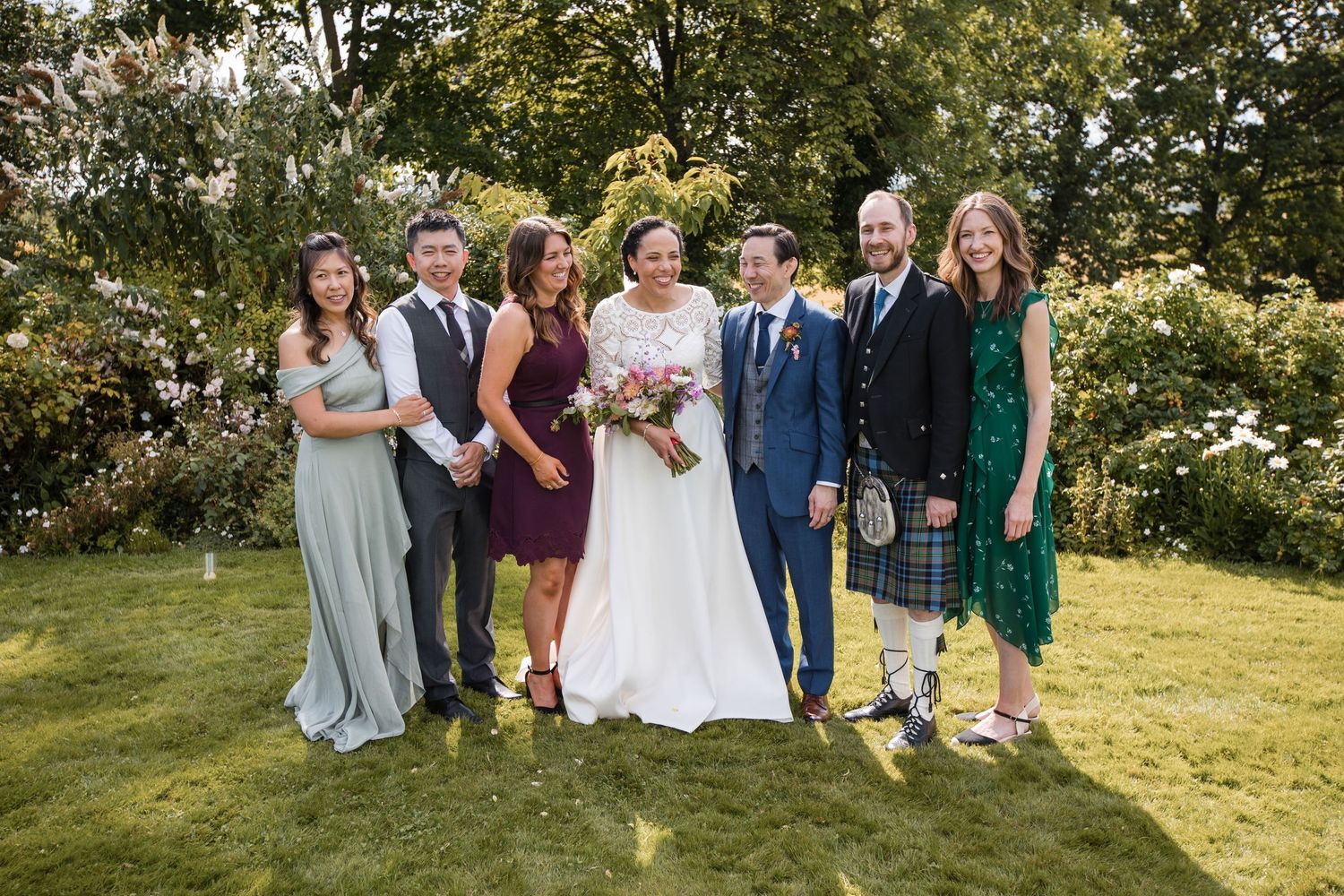 Wedding party members gathered on a green hillside for formal group portraits.
