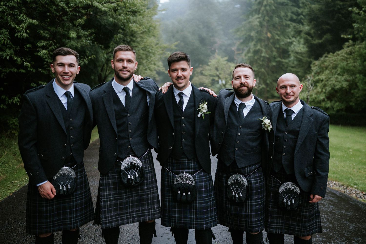 Groomsmen in traditional Scottish kilts pose together on a misty forest path.